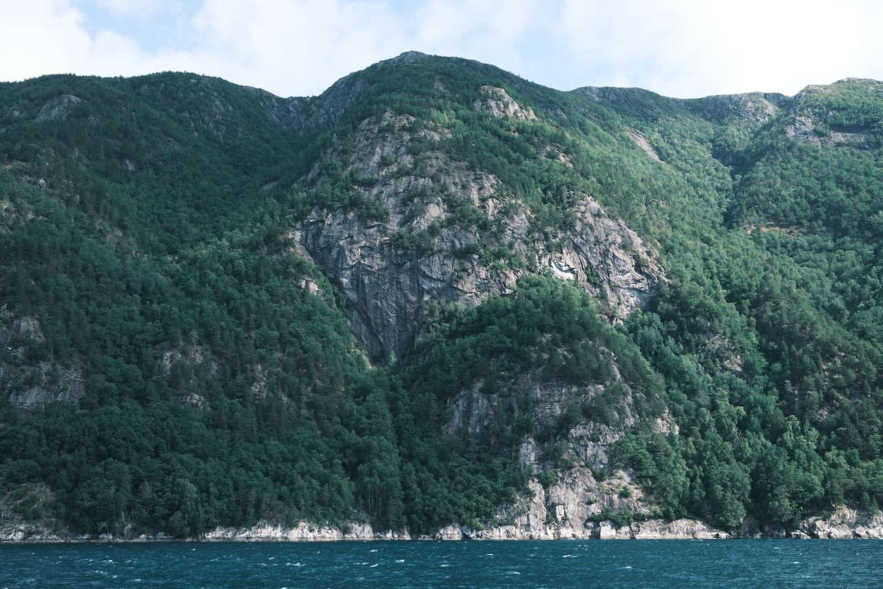 Auto-generated description: A large, forested mountain rises steeply from the edge of a body of water under a partly cloudy sky.