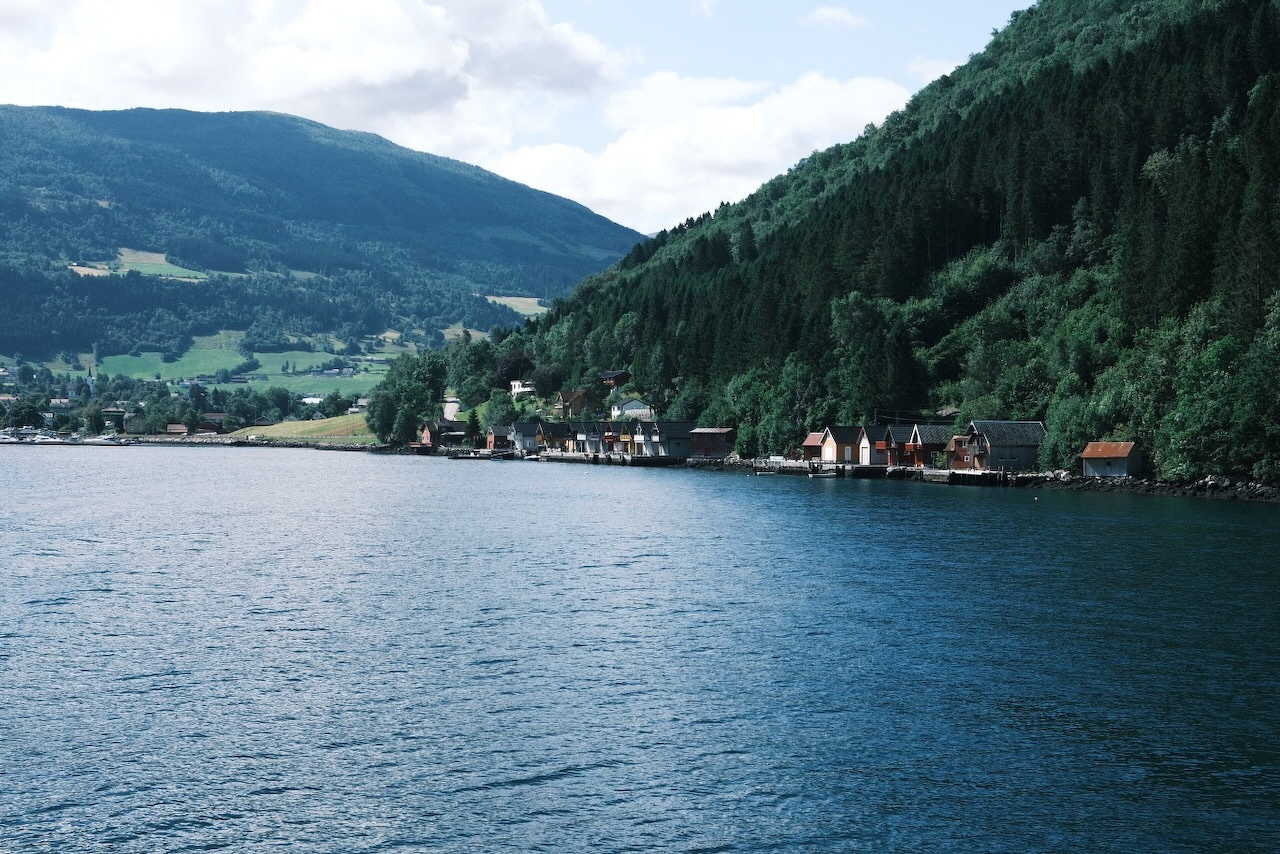 Auto-generated description: A peaceful lakeside village is nestled between dense green forests and gently rolling hills under a partly cloudy sky.