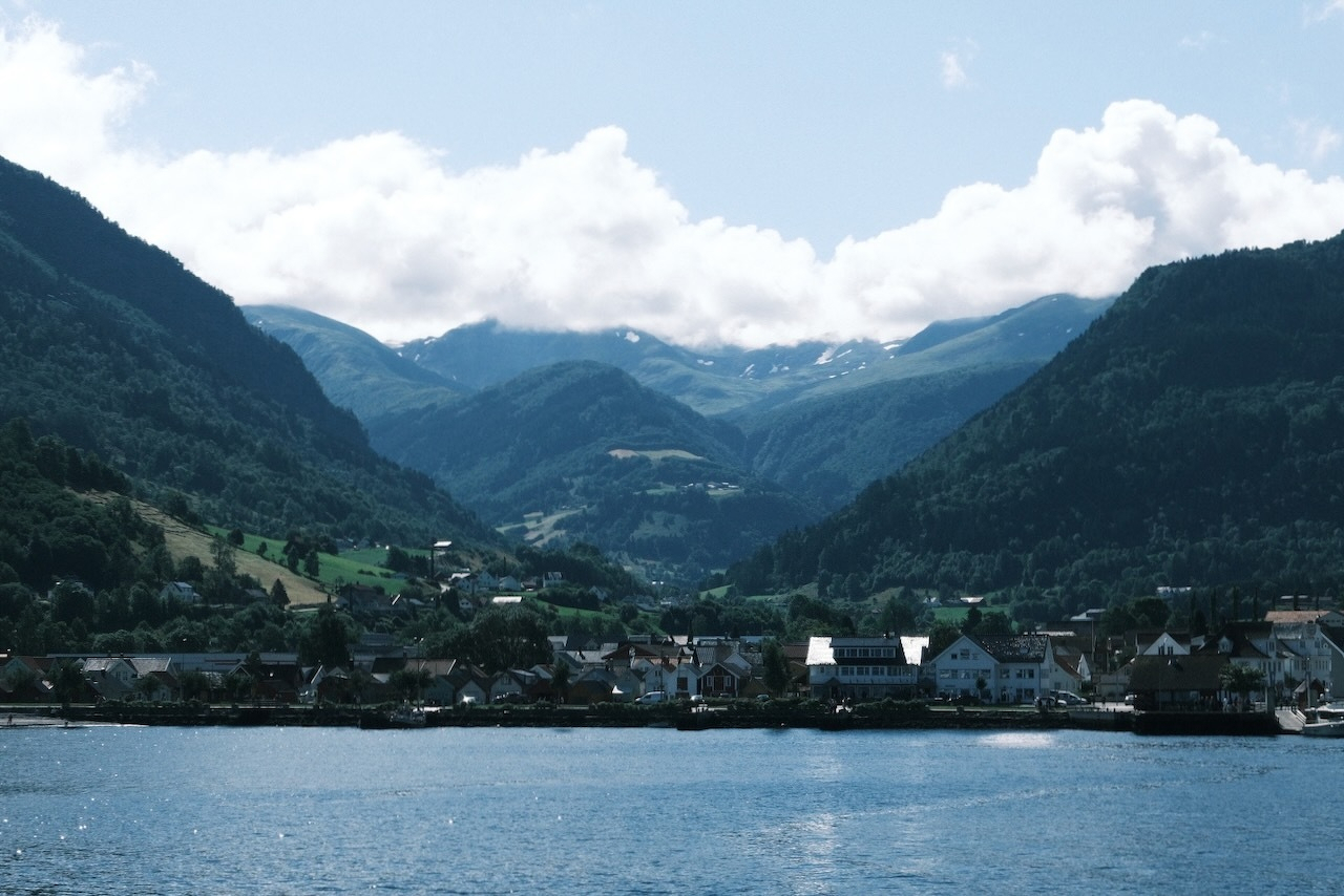 Auto-generated description: A small town is nestled at the base of green mountains under a partly cloudy sky, with a body of water in the foreground.
