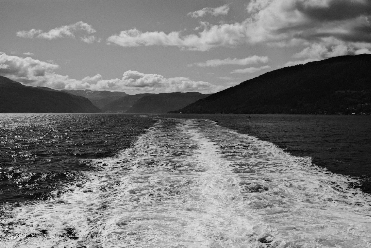Auto-generated description: A boat&rsquo;s wake stretches across a body of water surrounded by hills under a partly cloudy sky.