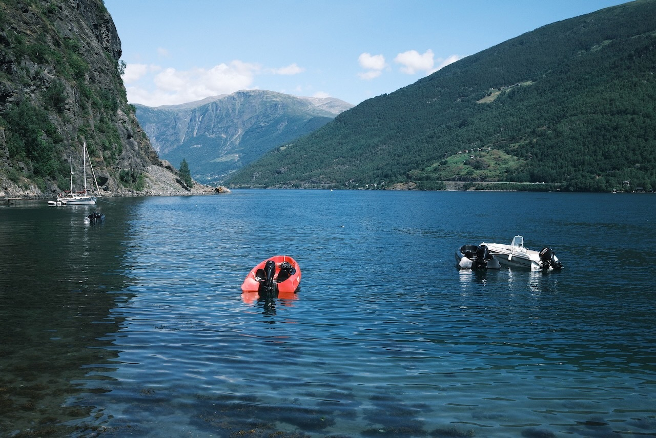 Auto-generated description: A person is floating on an orange inflatable ring in a calm body of water surrounded by lush green mountains and boats.