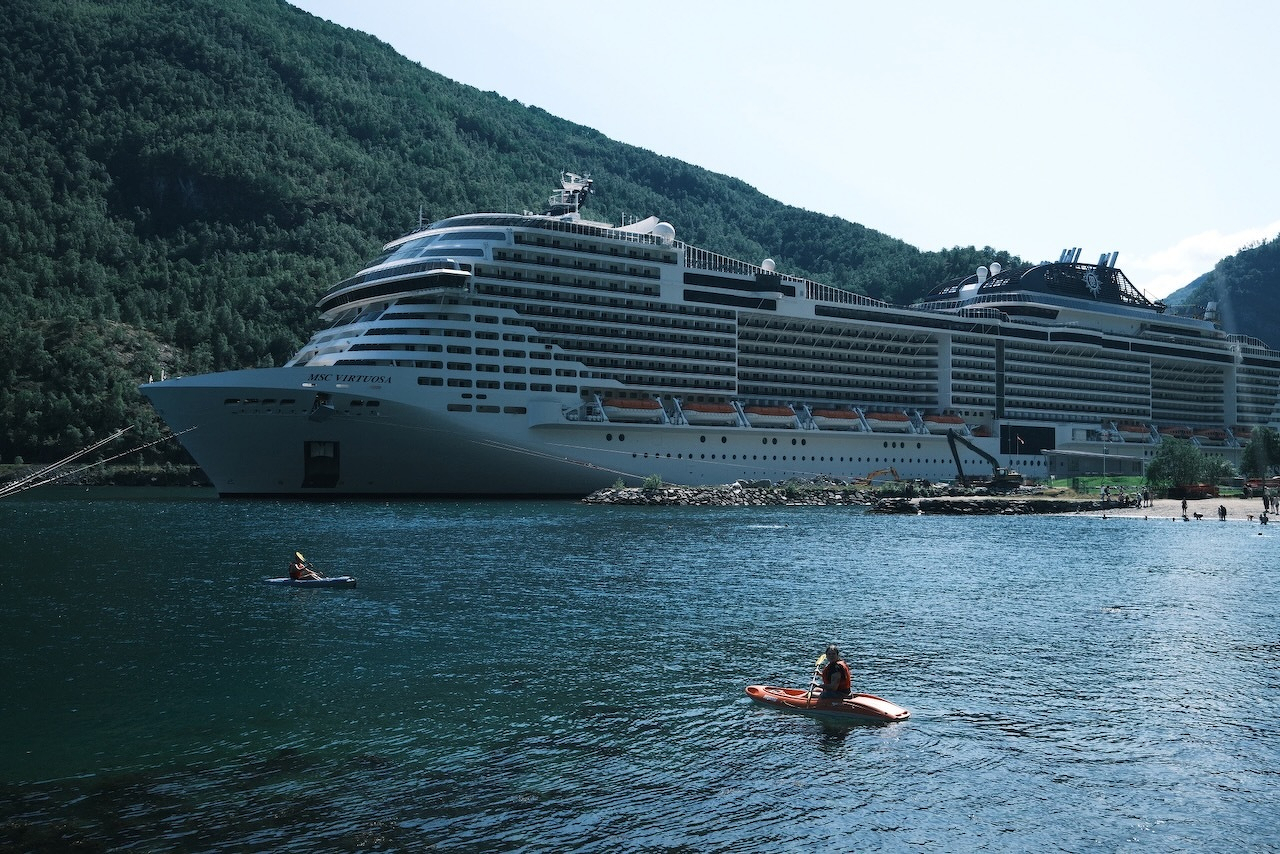 Auto-generated description: A large cruise ship is docked near a mountainous shoreline while two people paddle kayaks in the water nearby.