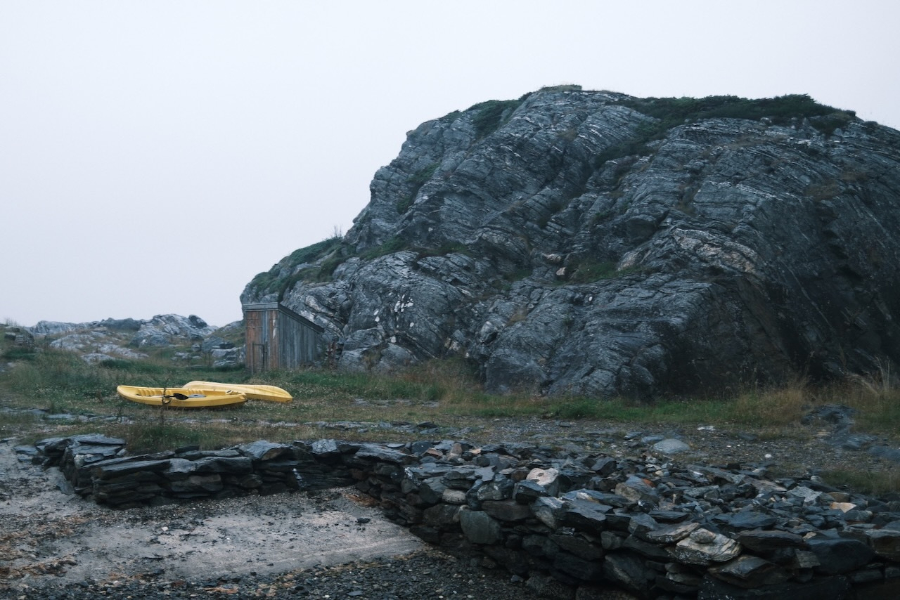 Auto-generated description: A rocky landscape features a large stone formation, a small wooden shed, a yellow kayak, and a low stone wall.