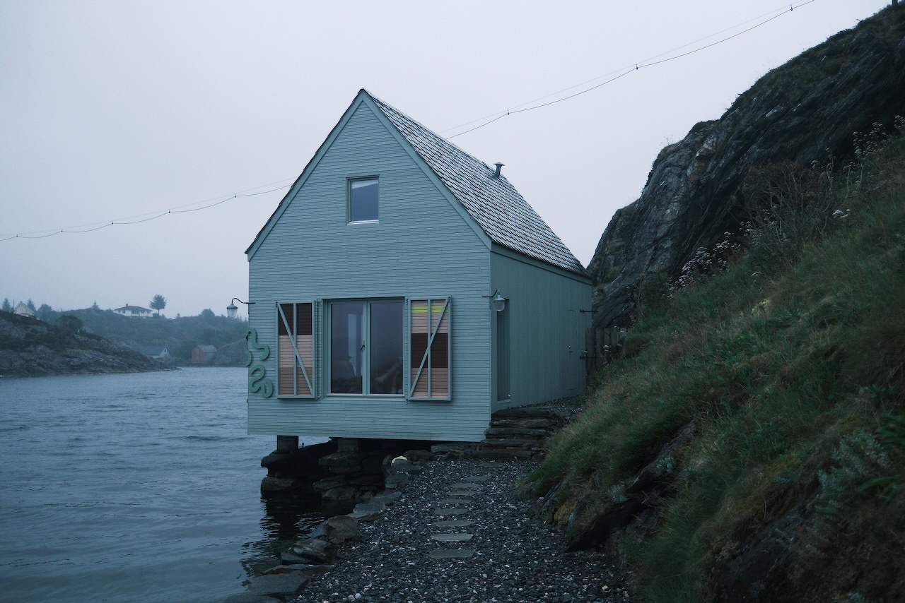 Auto-generated description: A small house on stilts is built at the edge of a rocky shore beside a steep hill, overlooking calm water under a cloudy sky.