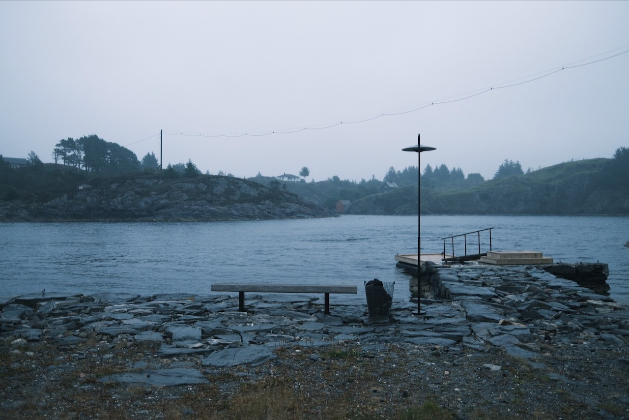 Auto-generated description: A calm waterfront scene features a stone pier with a bench and a tall, thin pole, set against a backdrop of misty hills and trees.