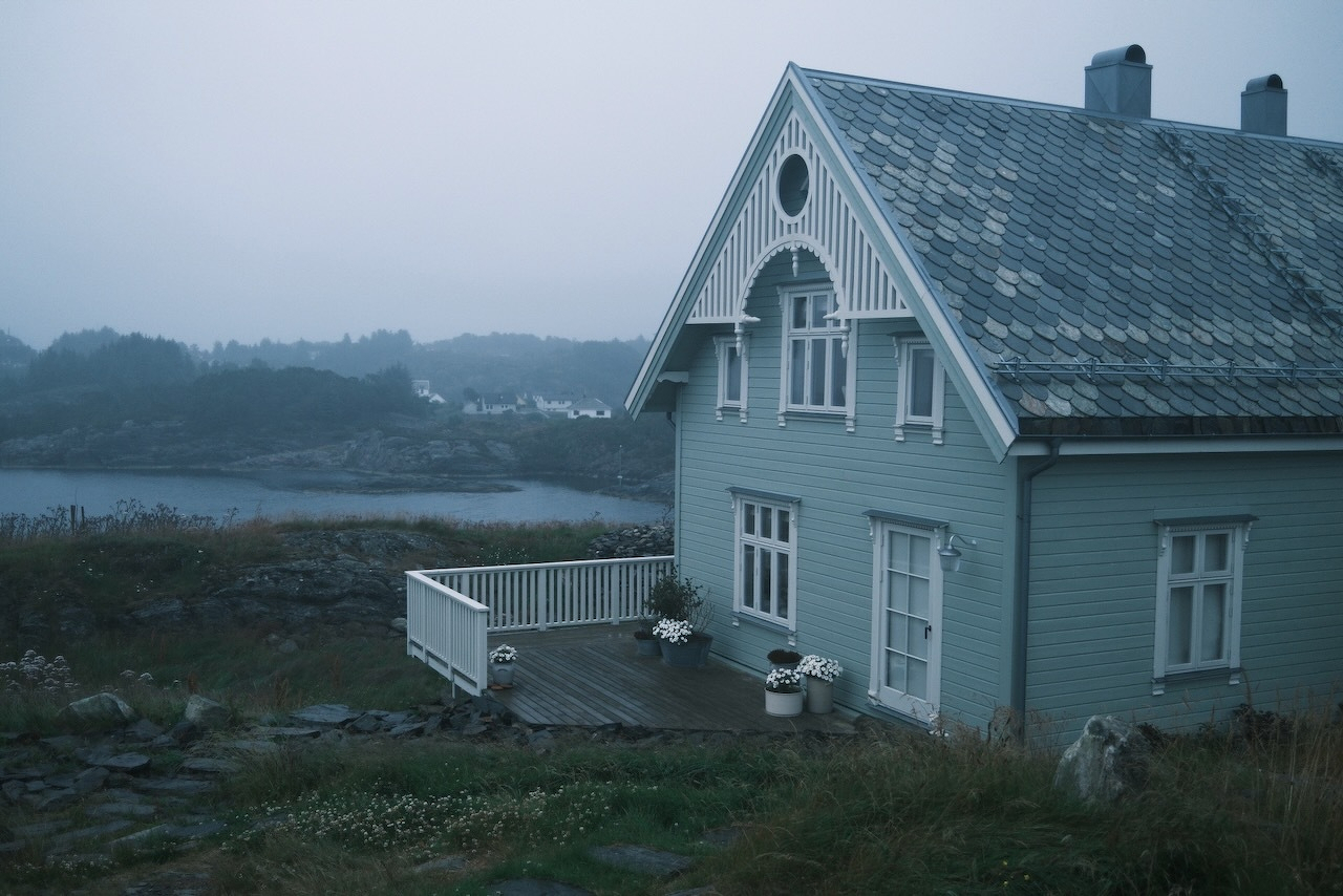 Auto-generated description: A gray house with a tiled roof sits on a grassy, rocky landscape near a body of water under a foggy sky.