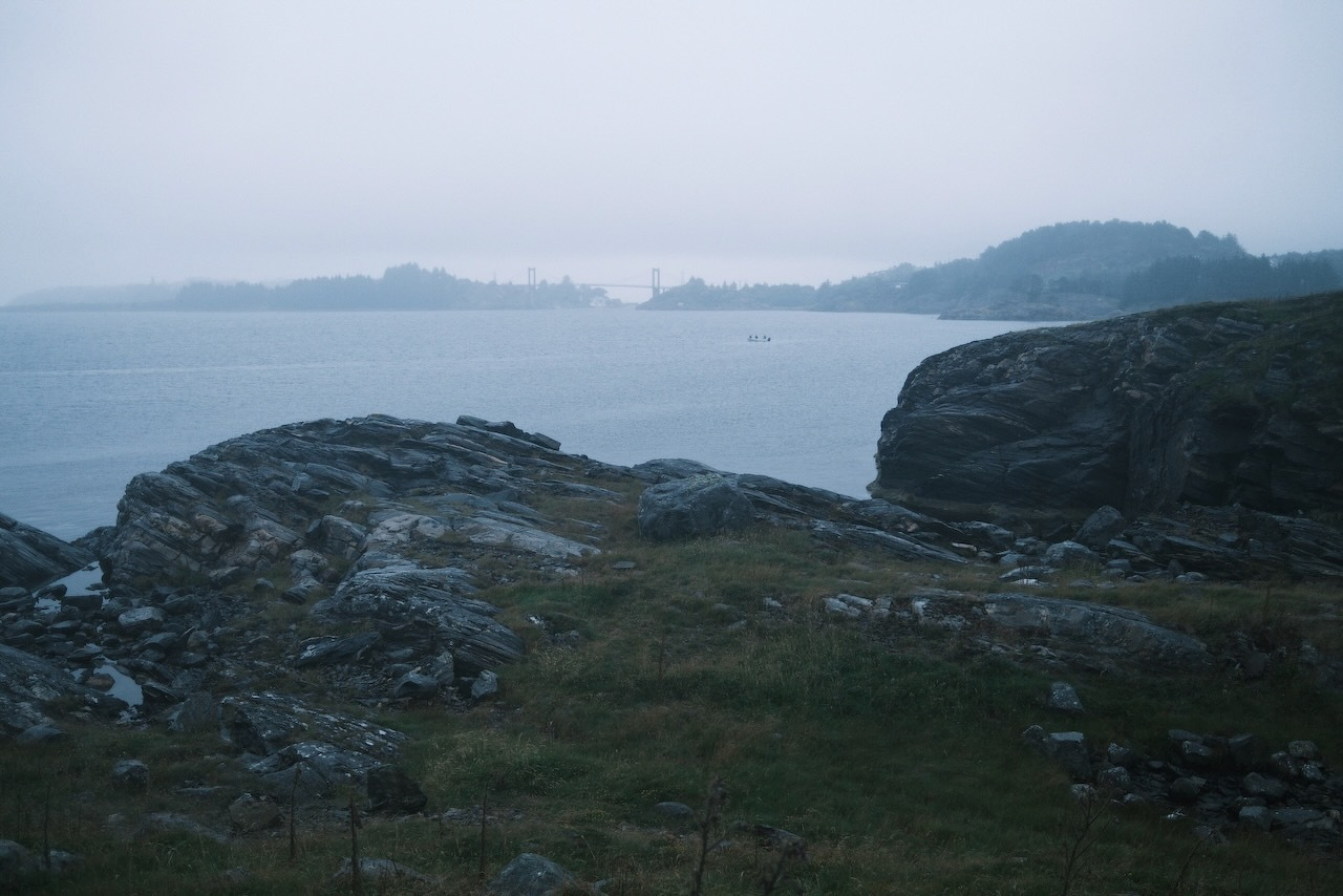 Auto-generated description: A rocky shoreline with patches of grass overlooks a calm body of water under a foggy or overcast sky.