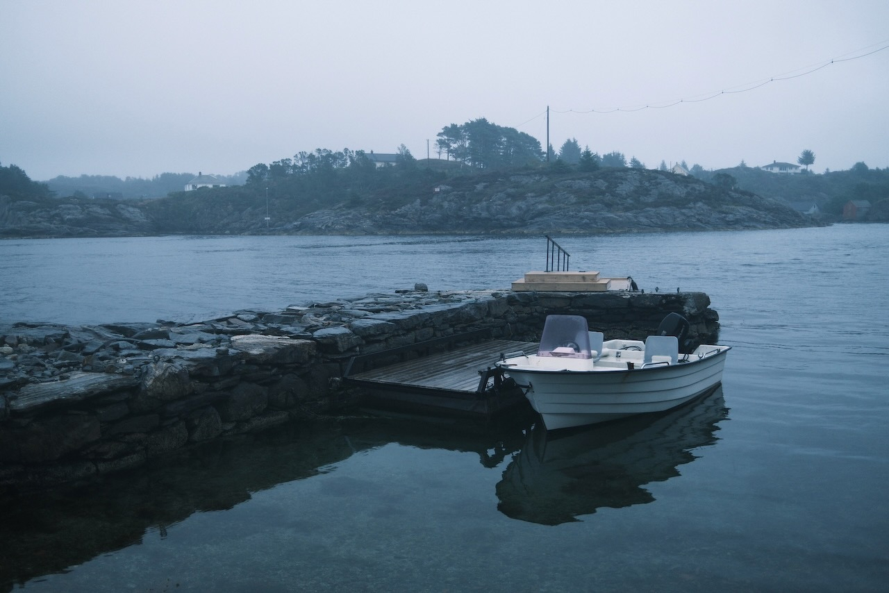 Auto-generated description: A small white boat is moored alongside a stone pier in calm waters with a misty, tree-covered island in the background.