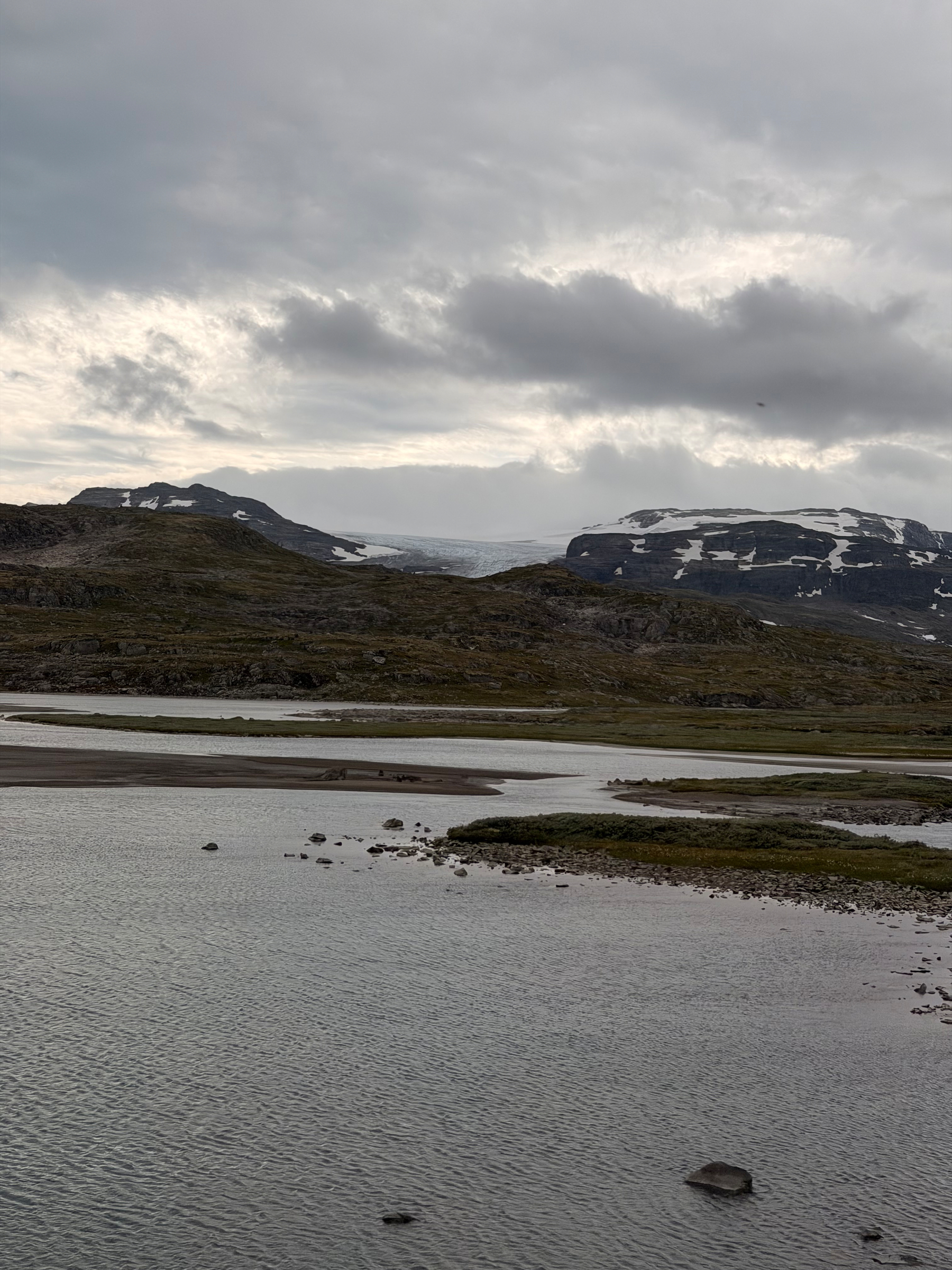 Auto-generated description: A calm river flows through a rocky, mountainous landscape under a cloudy sky with patches of snow on the peaks.