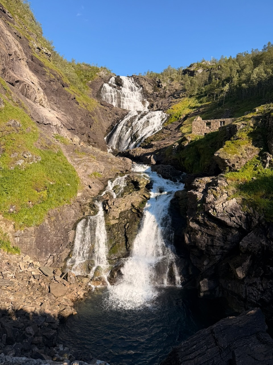Auto-generated description: A cascading waterfall flows down rocky cliffs surrounded by greenery under a clear blue sky, with an old stone structure visible on the right side.
