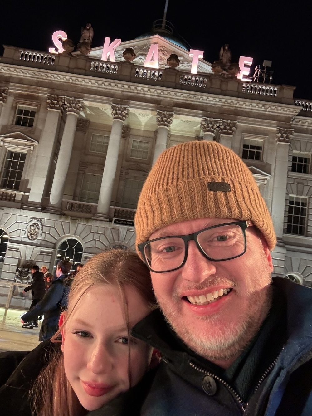 A man wearing glasses and a beanie poses with a young girl in front of a building illuminated with the word SKATE on top.