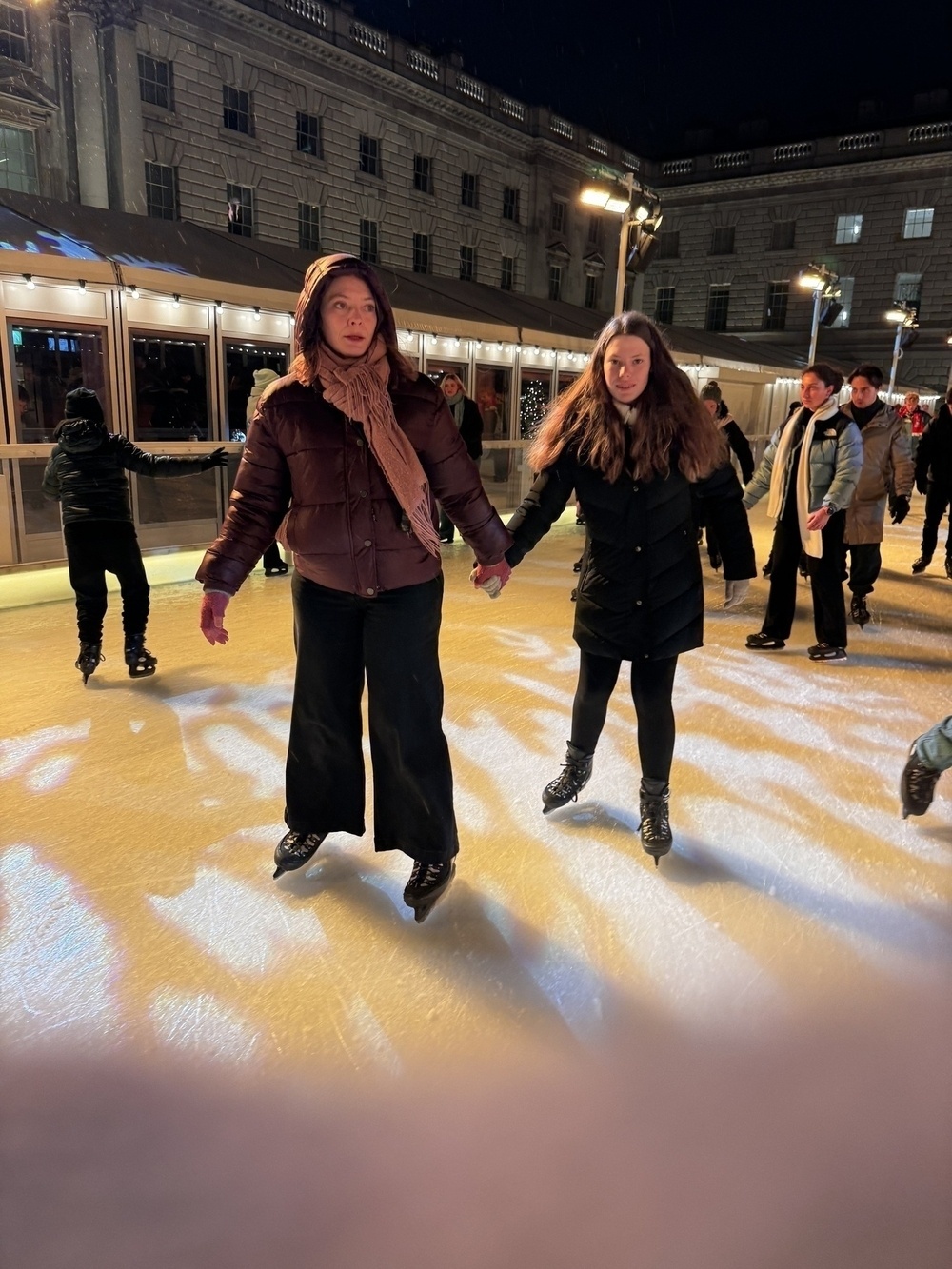 Two people are ice skating together on an outdoor rink at night, surrounded by other skaters.