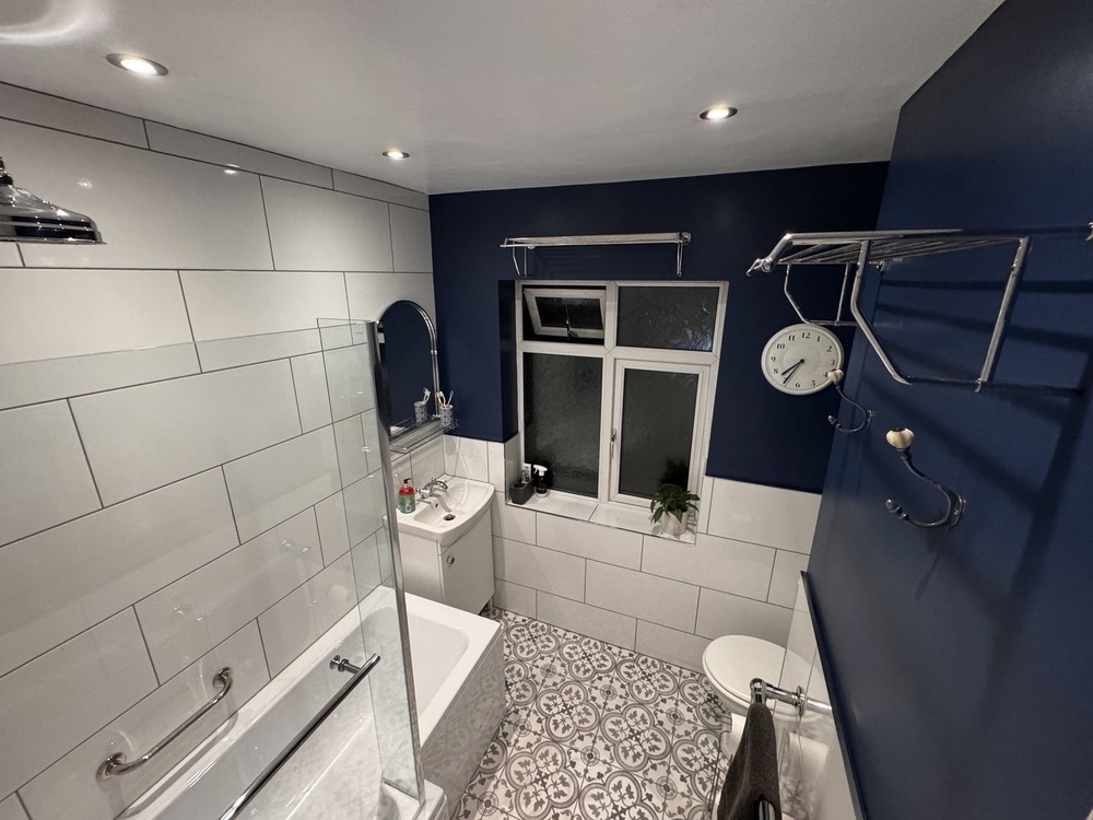 A modern bathroom features a combination of white and navy blue walls, a bathtub with a glass screen, patterned floor tiles, and a small window.