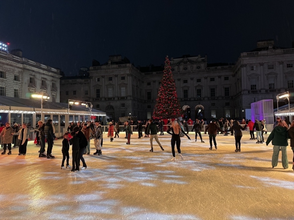 People are ice skating at an outdoor rink with a large decorated Christmas tree and historic buildings in the background.