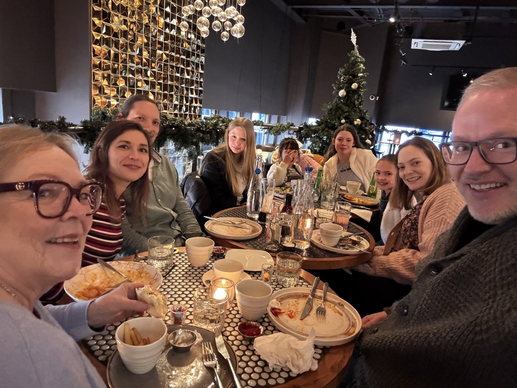 A group of people are sitting around a table enjoying a meal in a warmly decorated setting with a Christmas tree in the background.