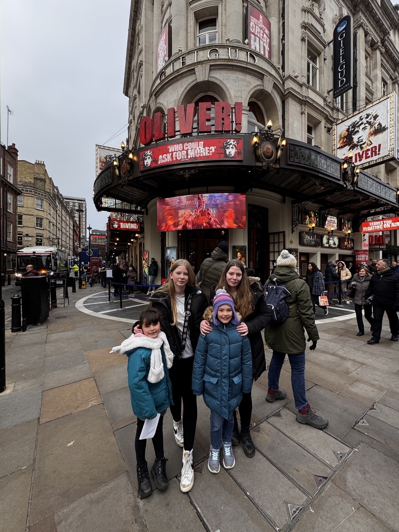 A group of people stands outside a theater displaying advertisements for a production of Oliver! in an urban setting.