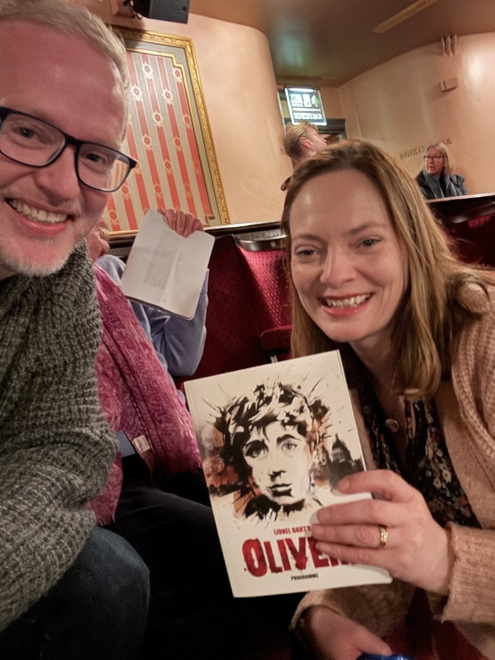 Two people are smiling in a theater, holding a program for Oliver!