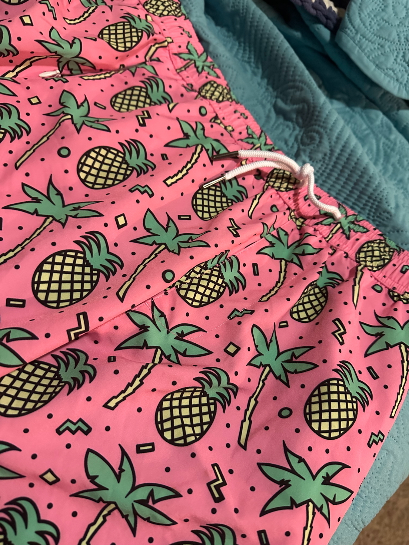 Pink shorts with a drawstring, featuring a playful pattern of green pineapples, palm trees, and abstract geometric shapes. The shorts are lying on a teal quilted surface.