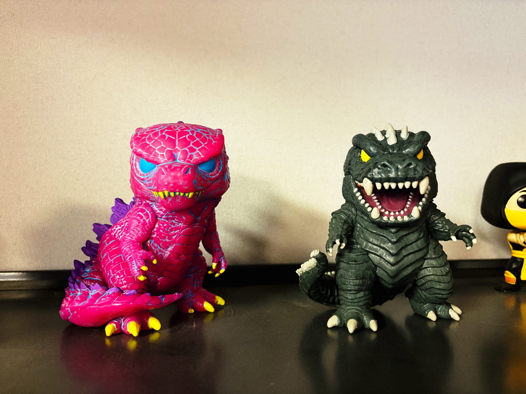 Two stylized Godzilla figures are shown. The left figure is bright pink with purple and yellow accents, while the right figure is classic dark gray with white and yellow details. They are placed on a black surface against a plain background.