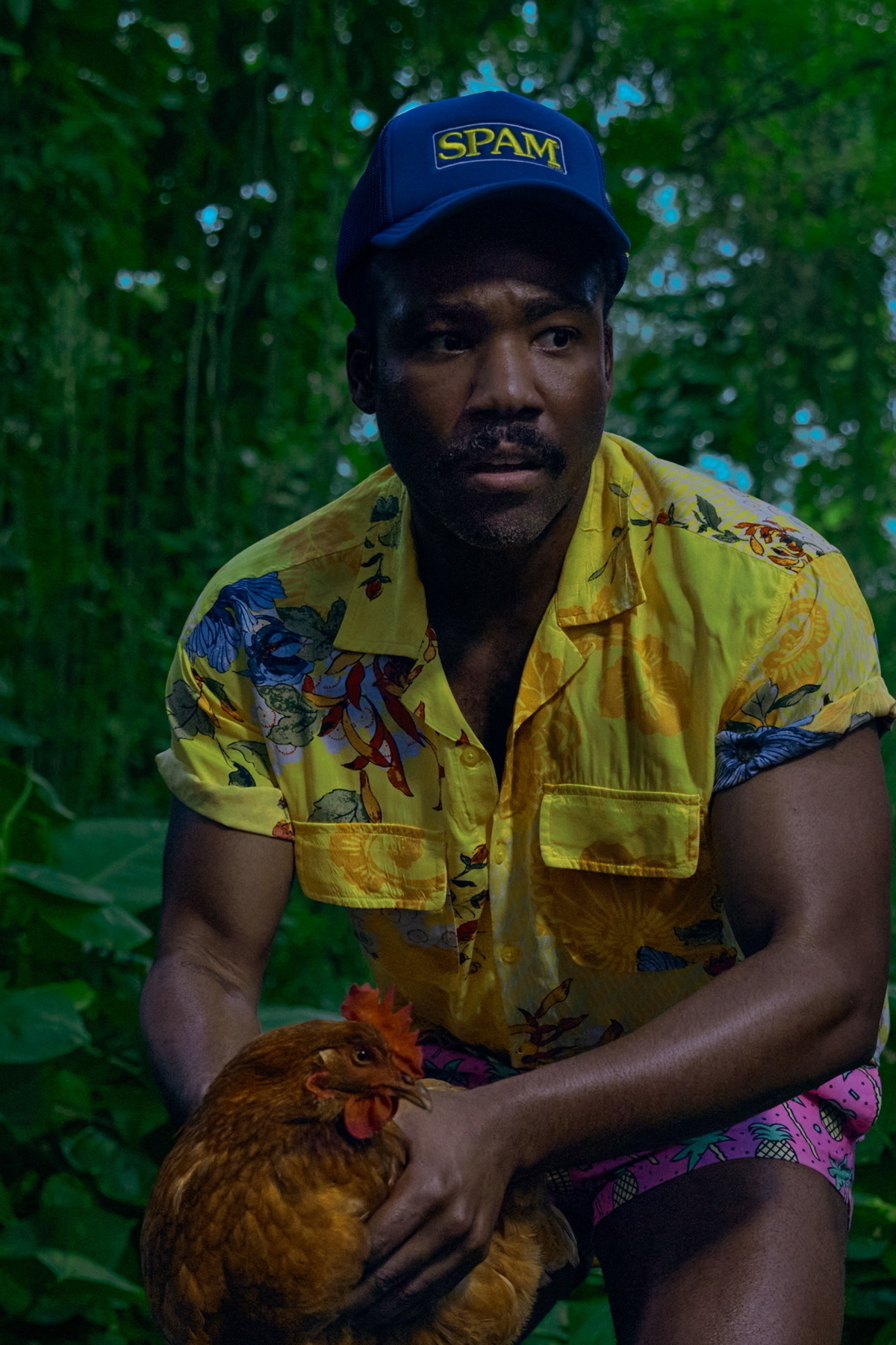 A man wearing a yellow floral shirt, shorts with a pineapple pattern, and a blue hat with the word “SPAM” is holding a brown chicken. The background is filled with lush green foliage.