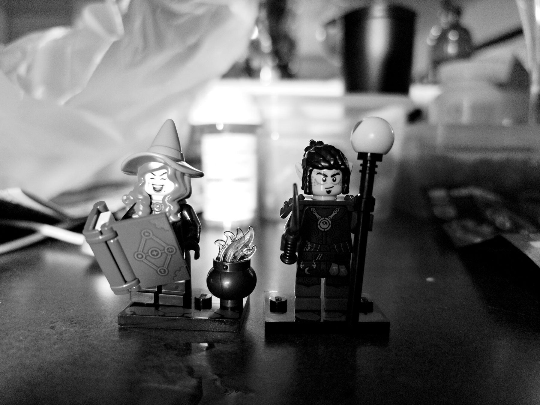 Two LEGO minifigures are displayed on a table. One is a wizard with a hat, holding a book, and the other is a warrior with a staff. A small cauldron with flames is between them. The background is blurred.