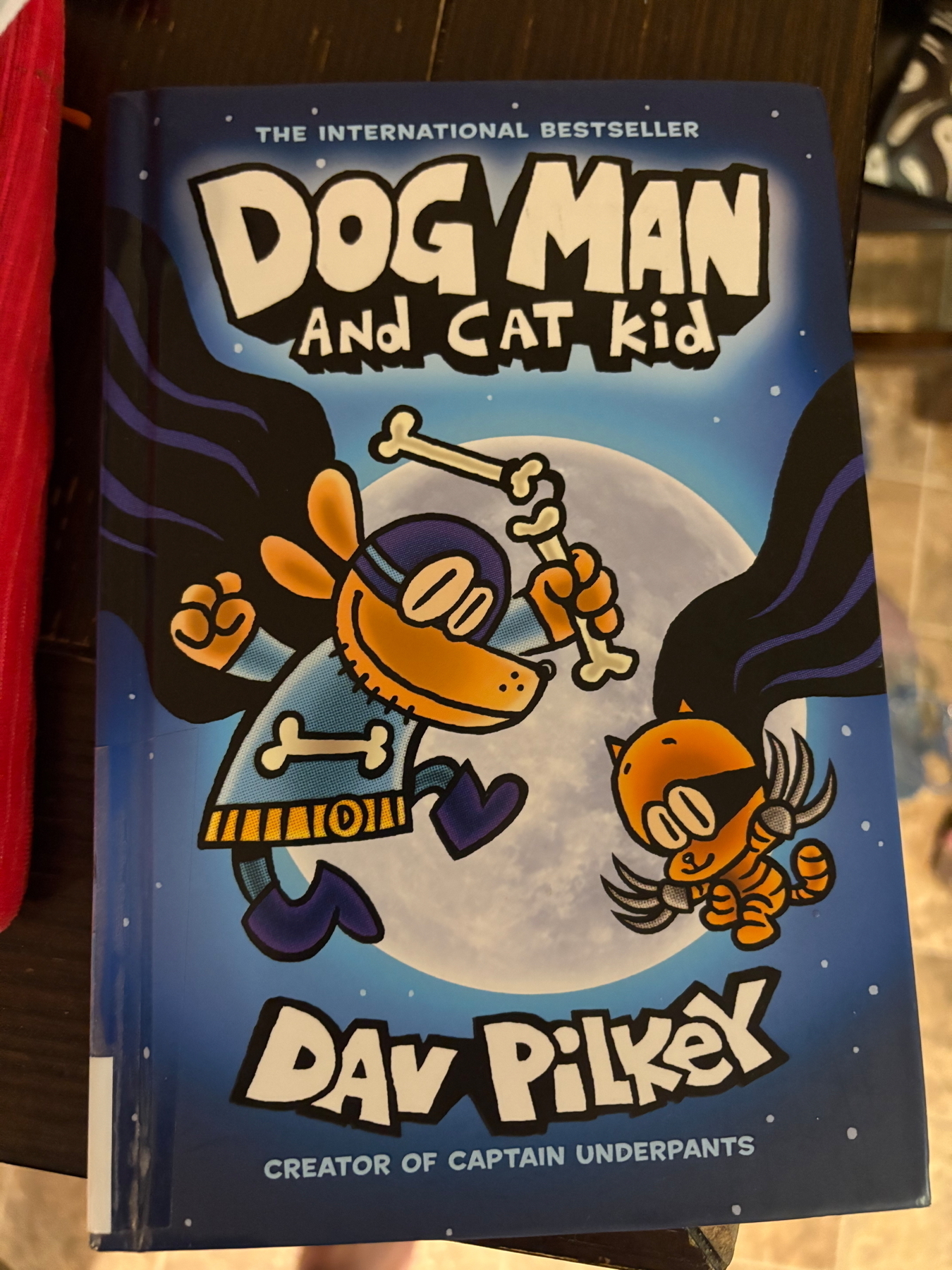 Cover of the book “Dog Man and Cat Kid” by Dav Pilkey. It features illustrations of Dog Man and Cat Kid in superhero costumes, flying against a full moon backdrop. The author’s name is prominently displayed at the bottom.