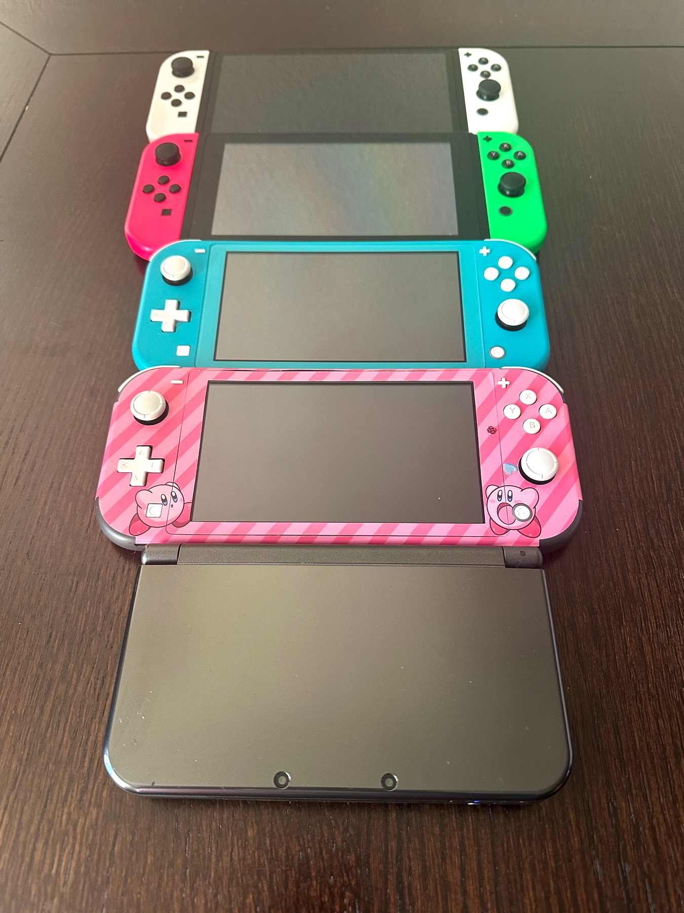 Four handheld gaming consoles on a wooden surface: a Nintendo 3DS at the bottom, a pink-themed Nintendo Switch Lite with character design above it, a blue Nintendo Switch Lite, and a standard Nintendo Switch with red and green Joy-Cons.