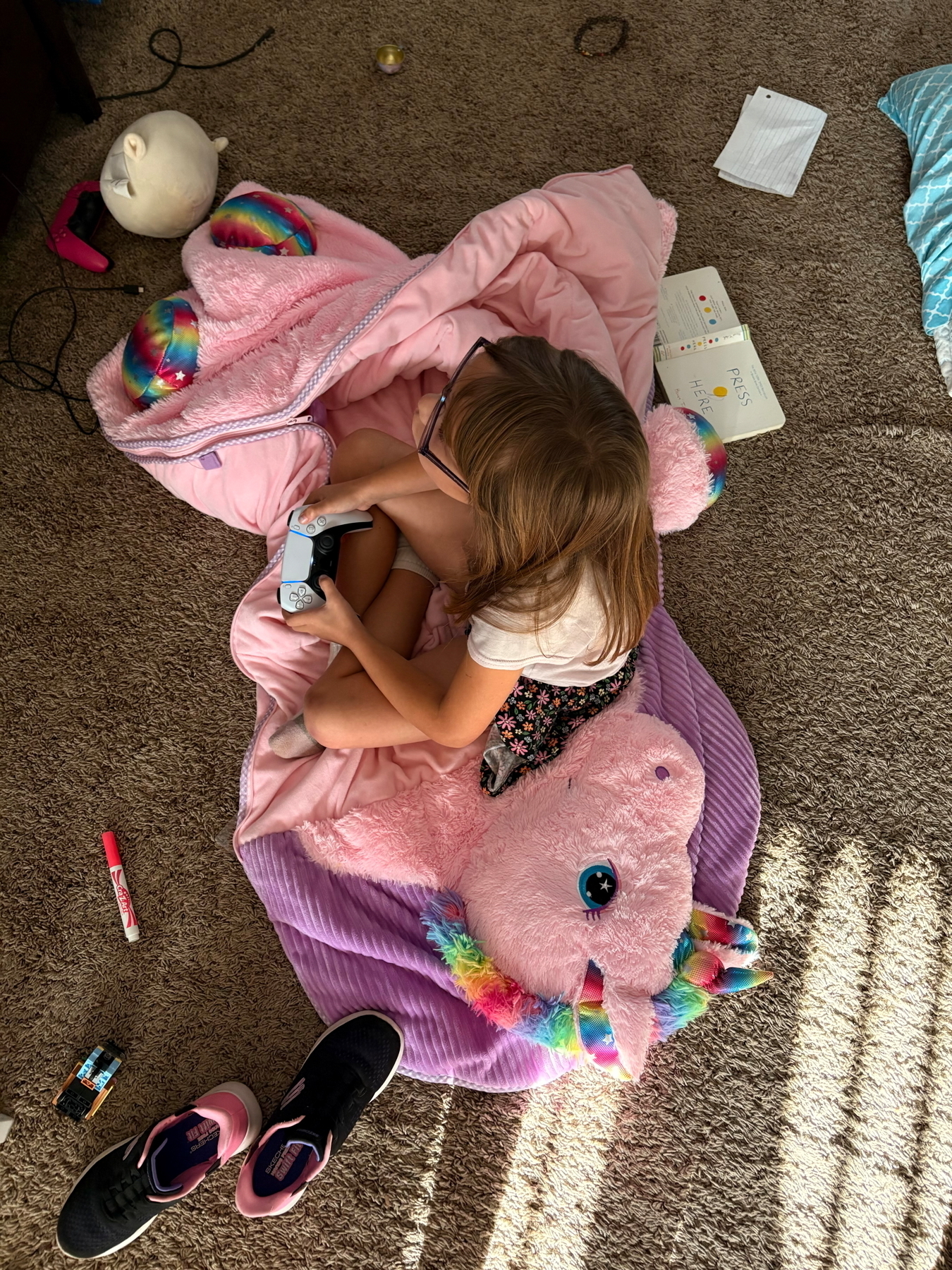 A child with glasses sitting on a pink and purple unicorn-themed blanket, playing a video game with a controller. Nearby are markers, a stuffed toy, gaming controllers, an open book, papers, and sneakers on a carpeted floor.