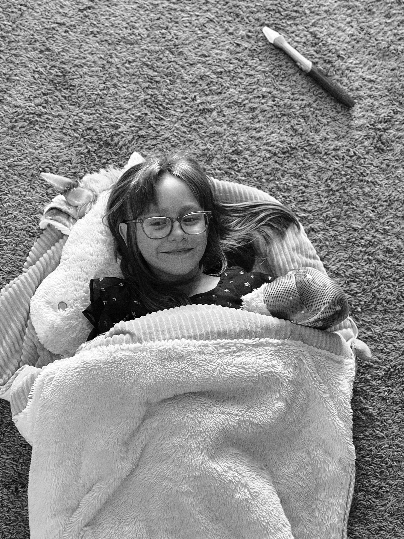 A black and white image of a young child lying on a carpet, wrapped in a fluffy blanket, with a plush toy beside them. The child is wearing glasses and smiling. A magic wand is on the carpet nearby.