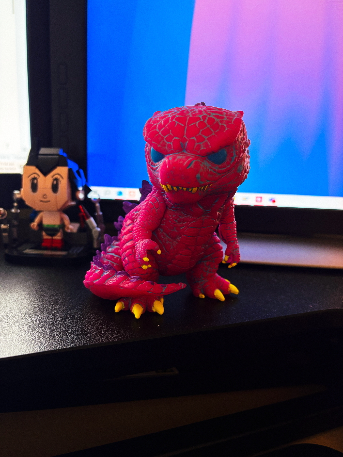 A brightly colored, pink and purple Godzilla toy with yellow claws is placed in front of a computer monitor. To the left, there is a small, square-headed toy figure.