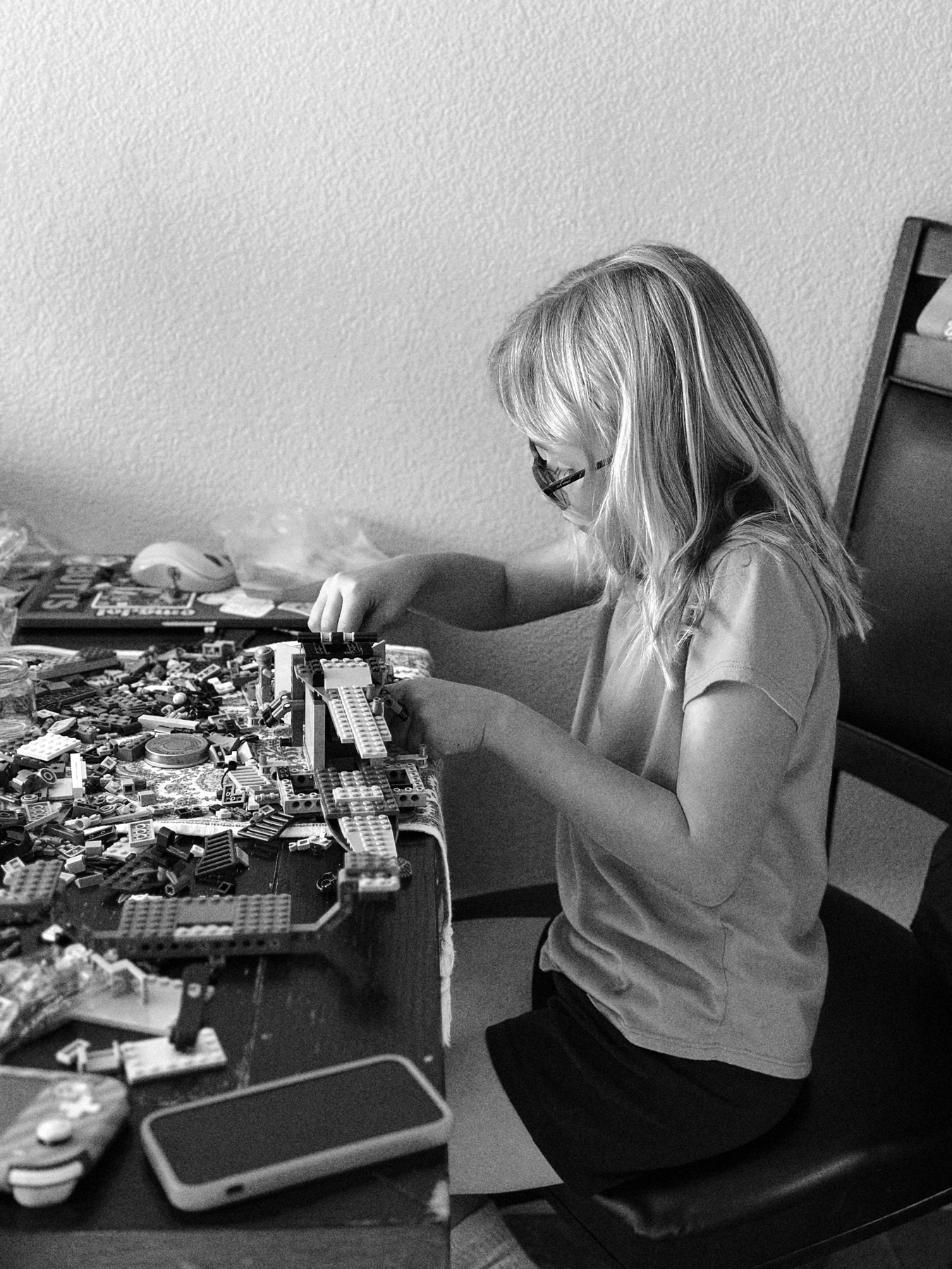 A black and white photo of a young person with glasses sitting at a table, focused on building with Lego bricks. Various Lego pieces and a smartphone are scattered on the table.