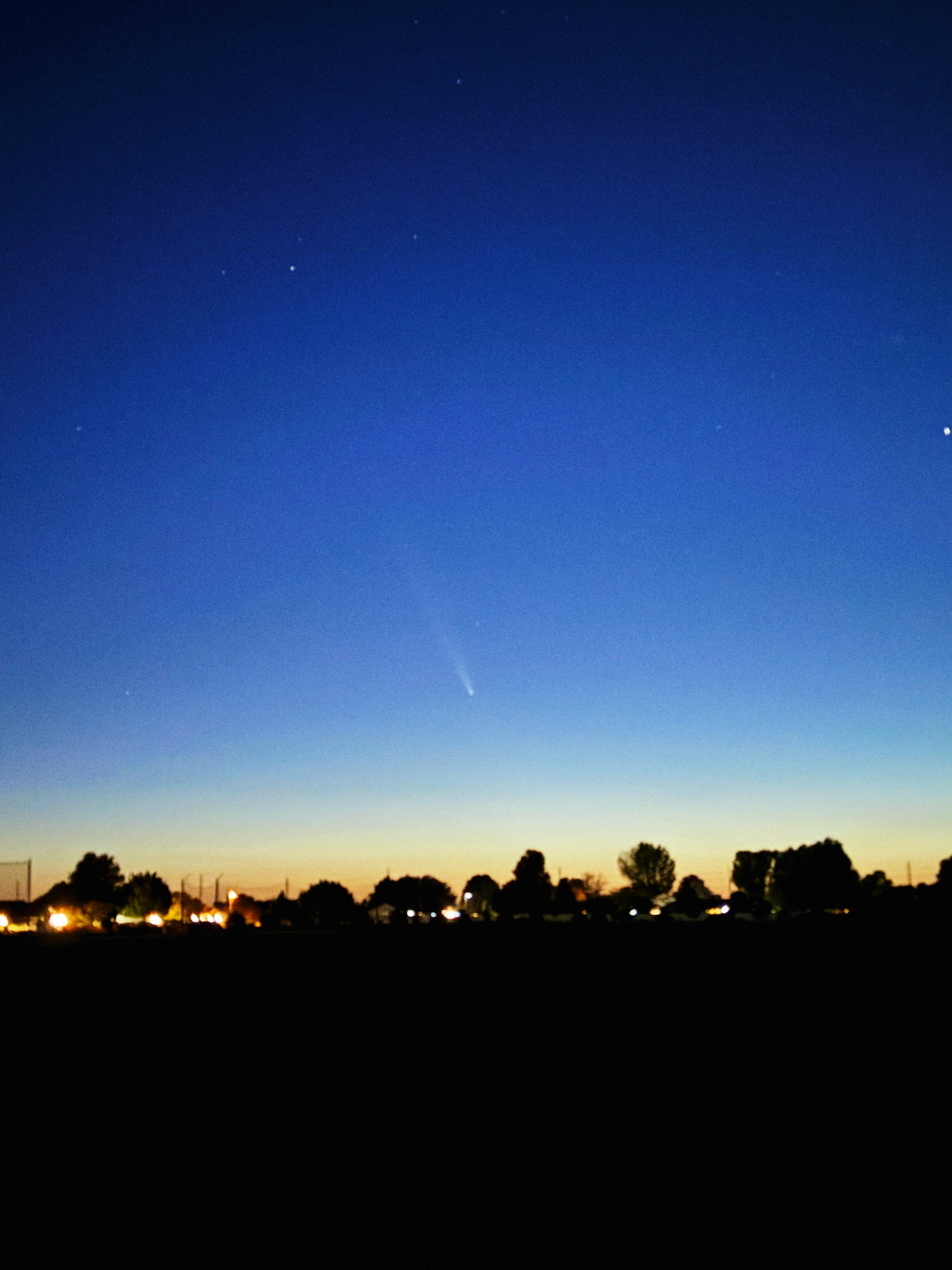 A twilight sky with a visible comet and stars, above a silhouetted landscape with trees and dimly lit houses.