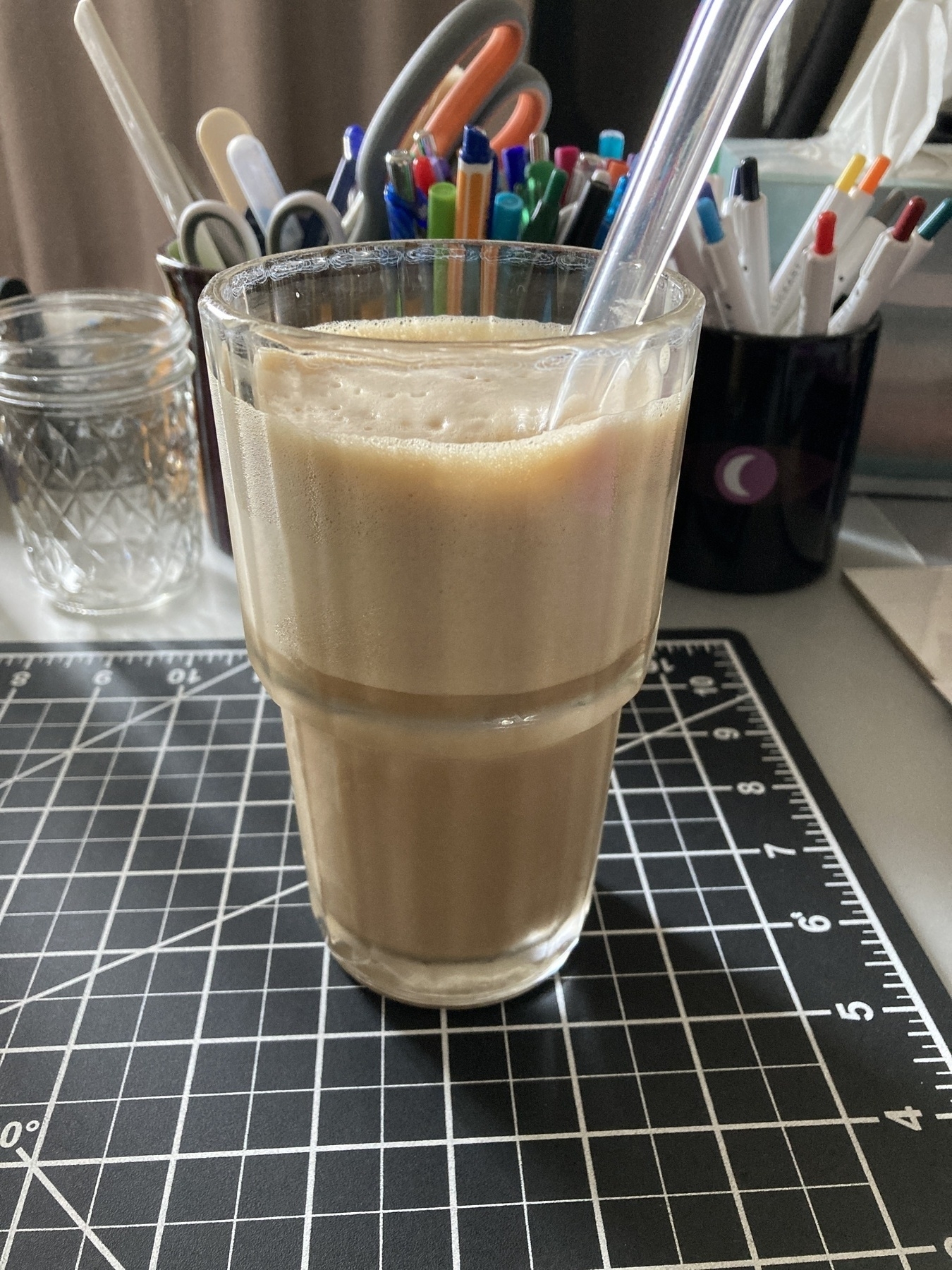 A glass of iced coffee with a straw sits on a black cutting mat near jars of colorful pens and markers.