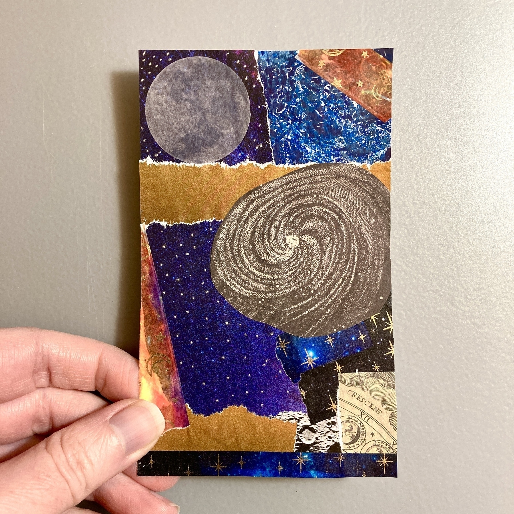 A hand holds a small collage made up of outer-space related imagery: a full moon and a galaxy against a background of blue and purple night skies. Orange stripes are in contrast to the blues and purples.