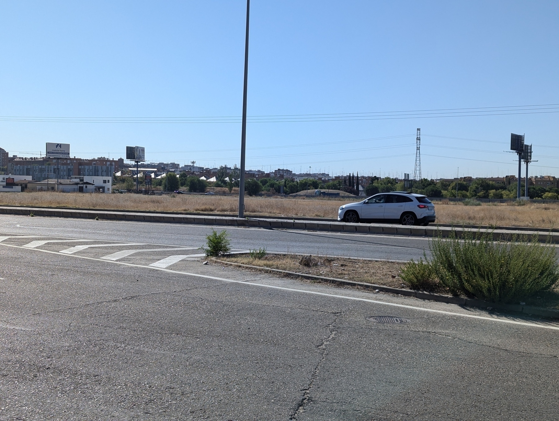 Auto-generated description: A white car drives along a road beside an empty field, with buildings and power lines in the background.