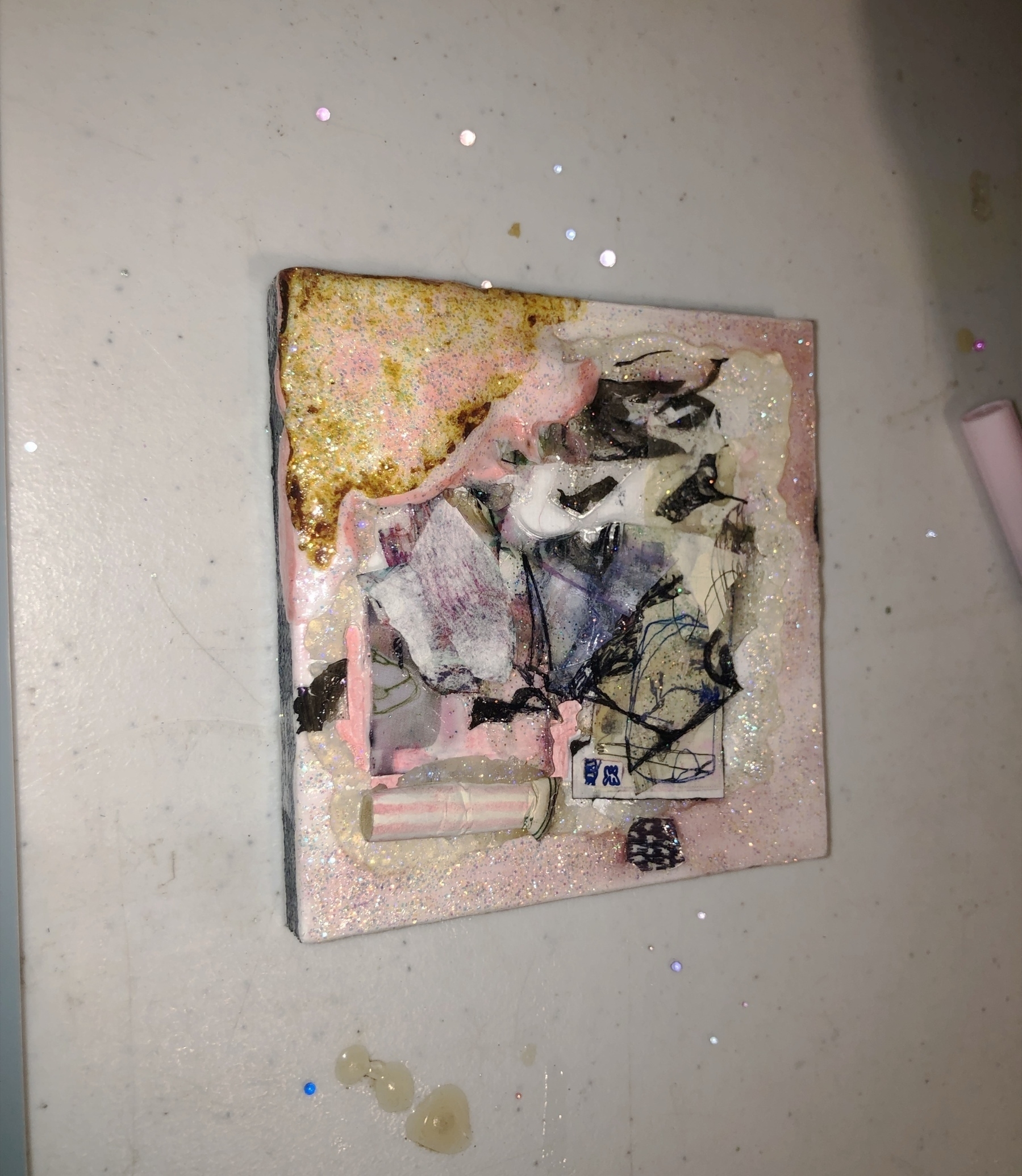 Auto-generated description: A small, abstract mixed-media artwork features various textures and colors, including pink, black, and gold, on a flat surface.