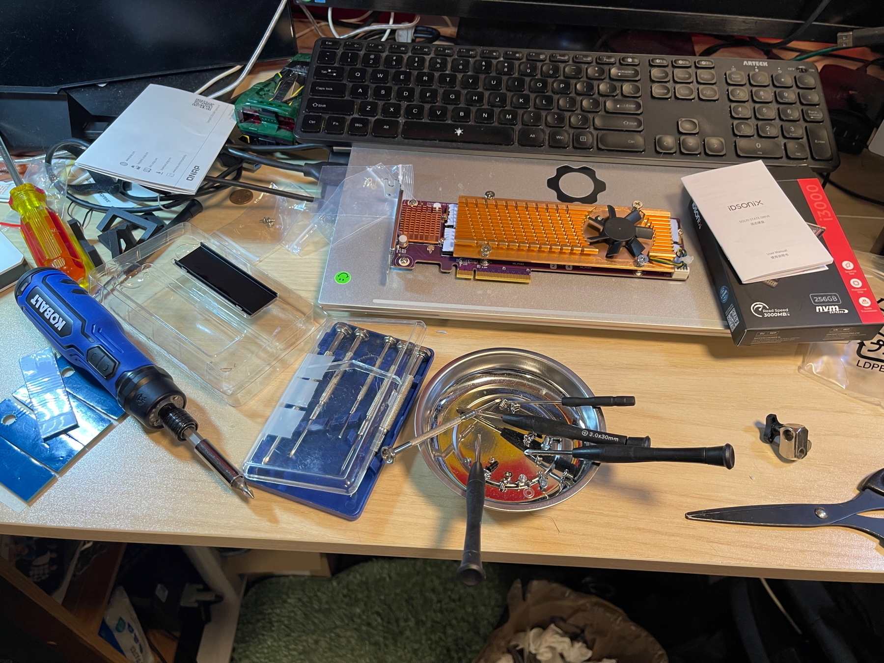 A cluttered desk with various tools, electronic components, a keyboard, and several small boxes organized around a central workspace.