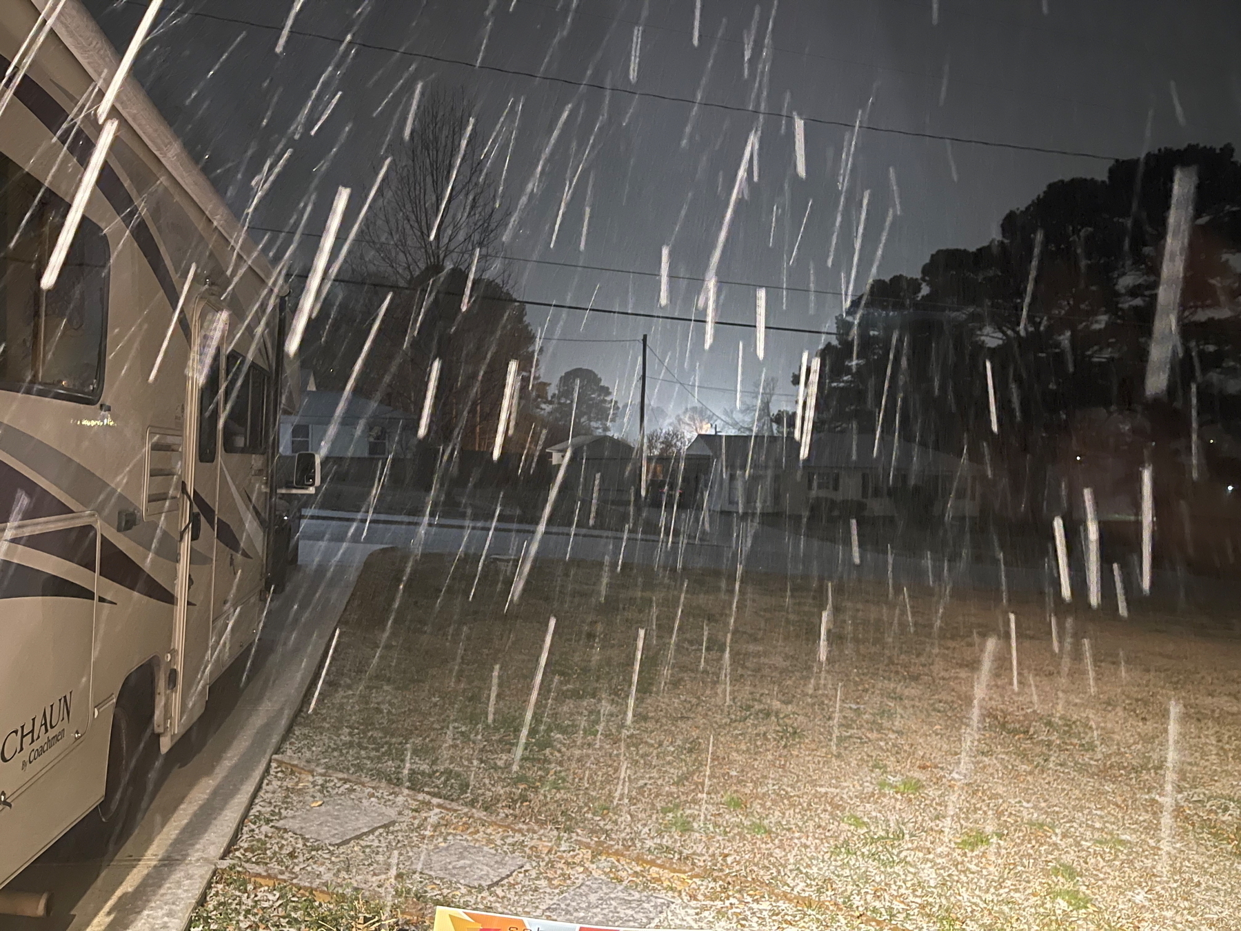 Auto-generated description: Snow falls heavily at night in a suburban neighborhood with a motorhome parked nearby.