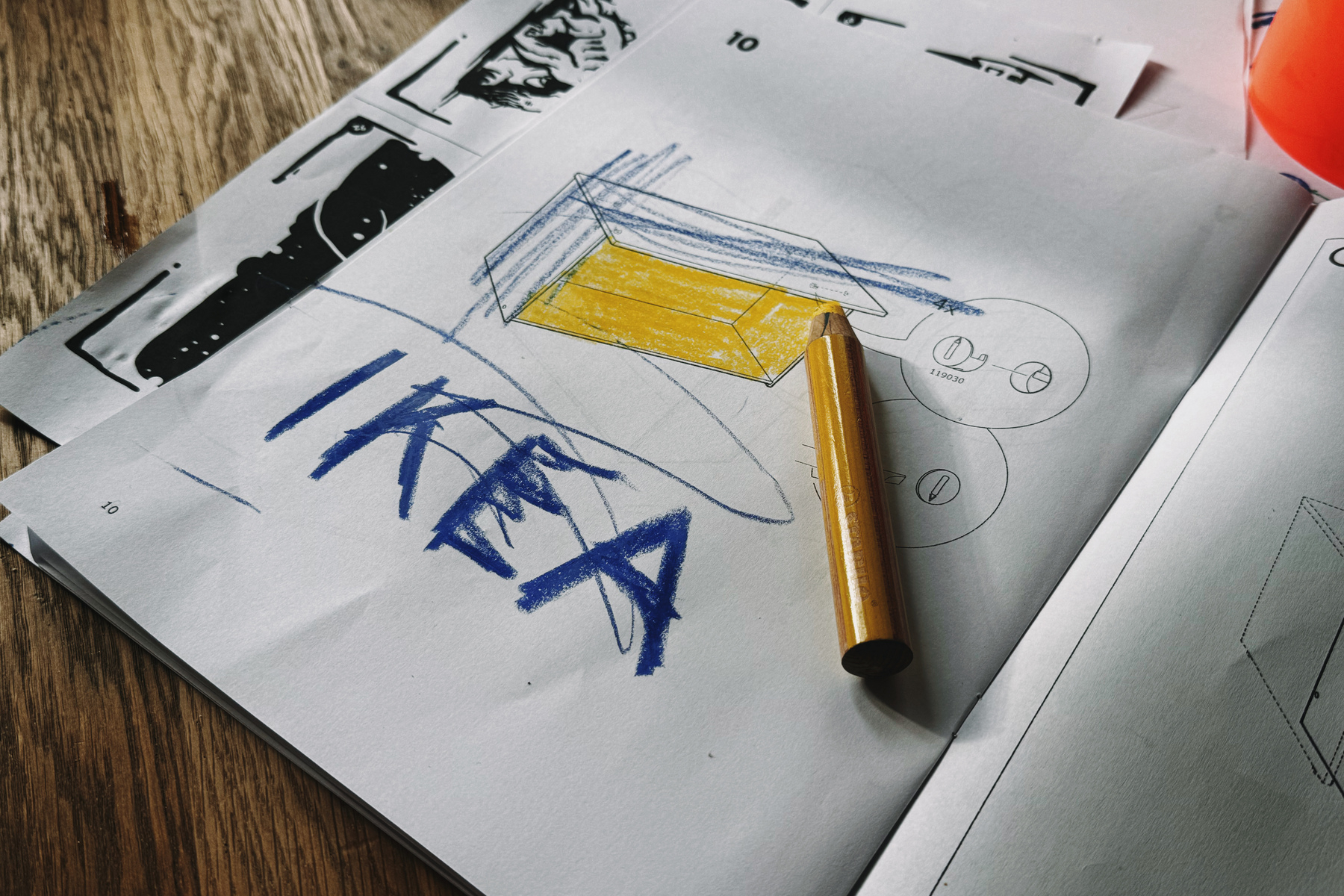 A pile of papers with illustrations is topped by a yellow crayon and the word "IKEA" hand-drawn in large blue letters.