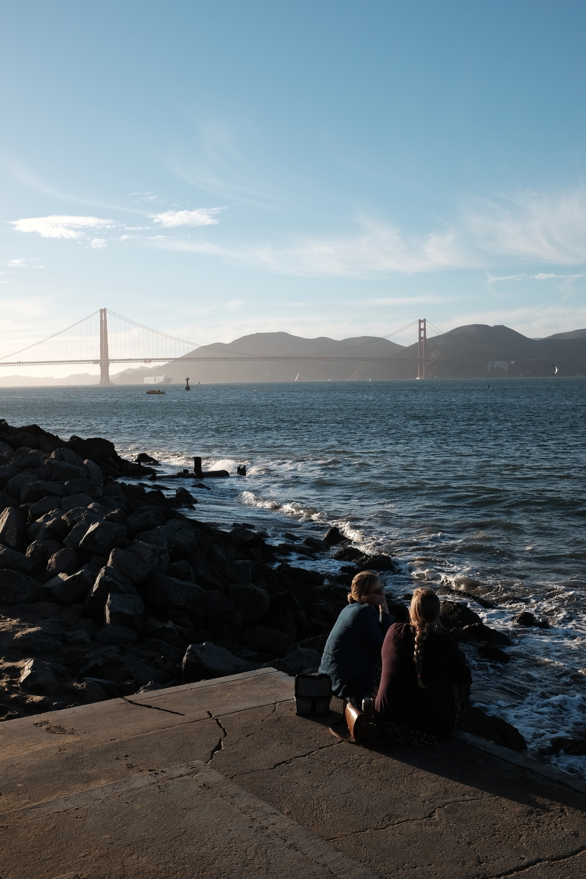 Two people sit on a rocky shoreline with a view of the Golden Gate Bridge in the background under a clear blue sky.