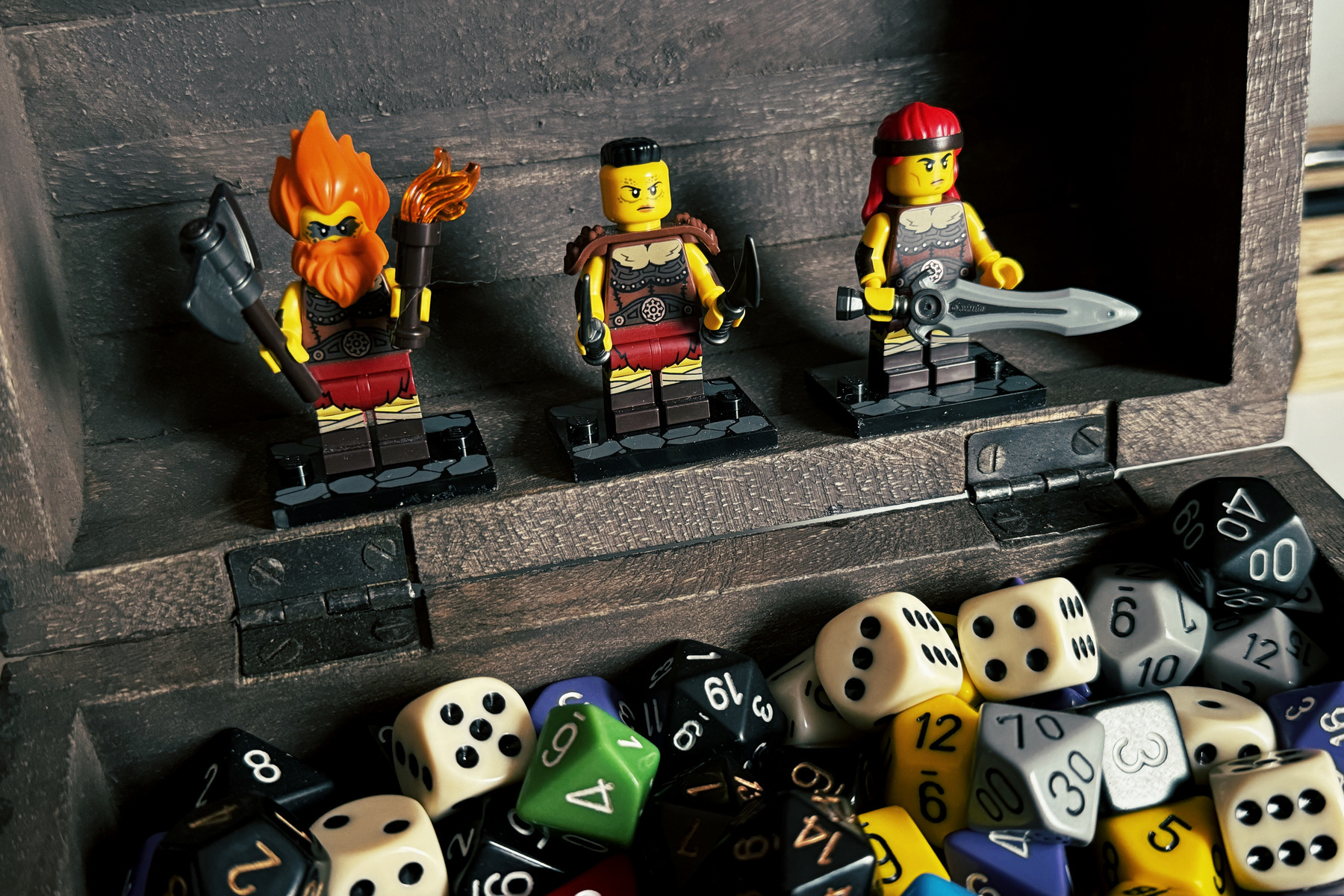 A group of three toy figures resembling warriors is displayed inside a wooden box filled with various colorful dice.