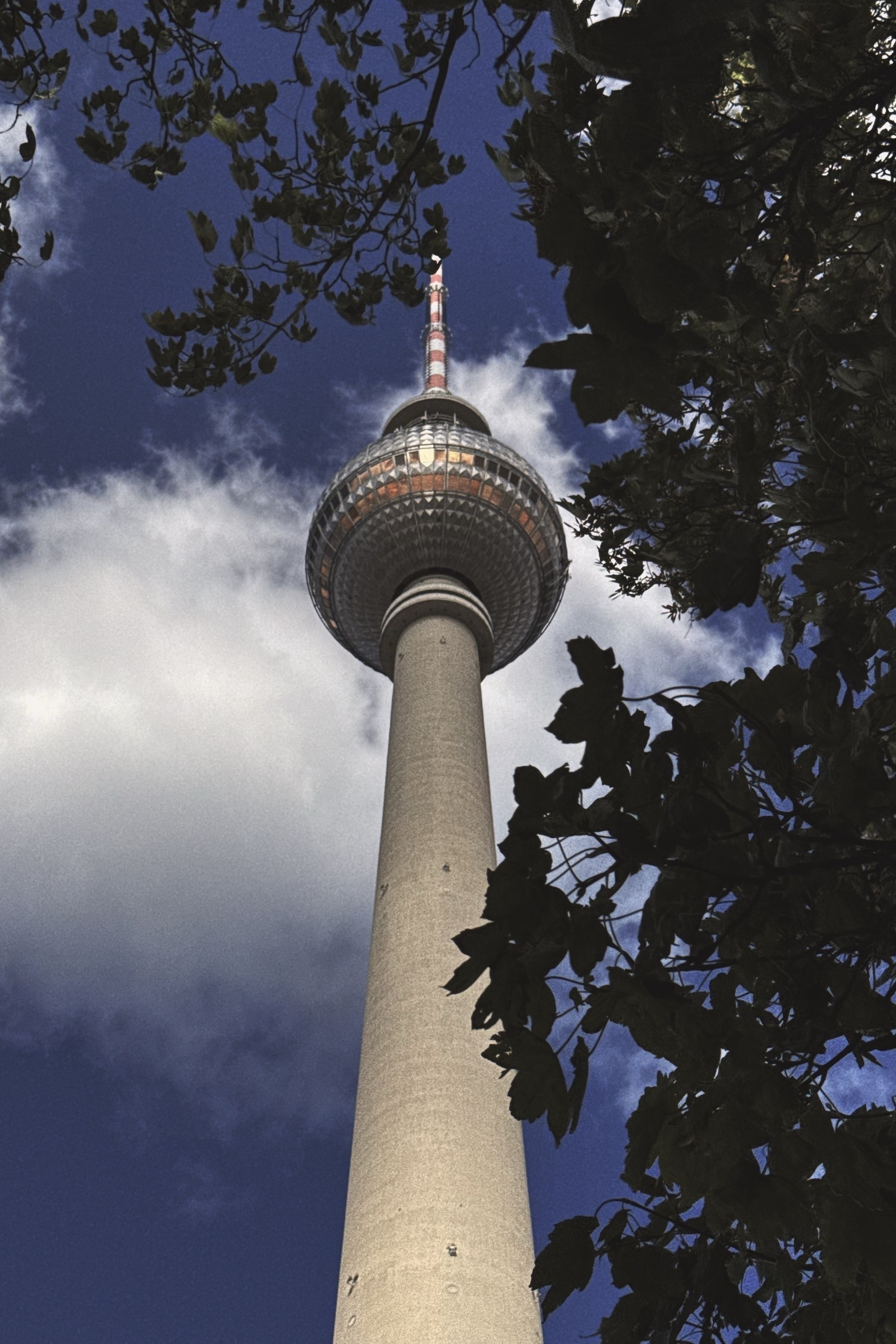 A tall, cylindrical tower with a spherical observation deck is framed by leafy branches against a sky with clouds.