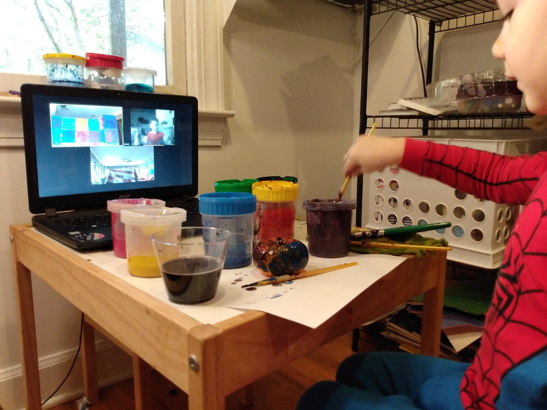 A white boy in Spider-Man pajamas paints while a laptop in front of him displays a video call with other young children.