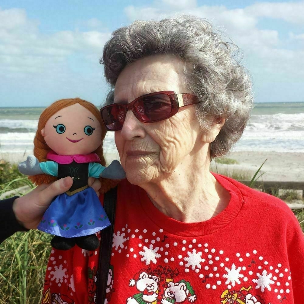 A doll of Anna from Frozen sits on the shoulder of an elderly white woman. They are in front of a beach. 