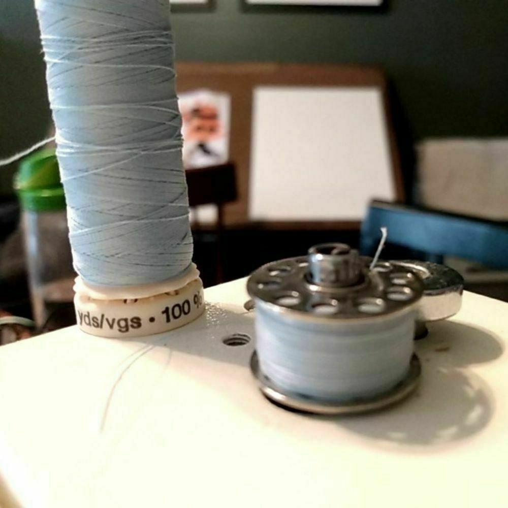 A bobbin with light blue thread sits atop a sewing machine. A spool of thread the same color sits behind it.
