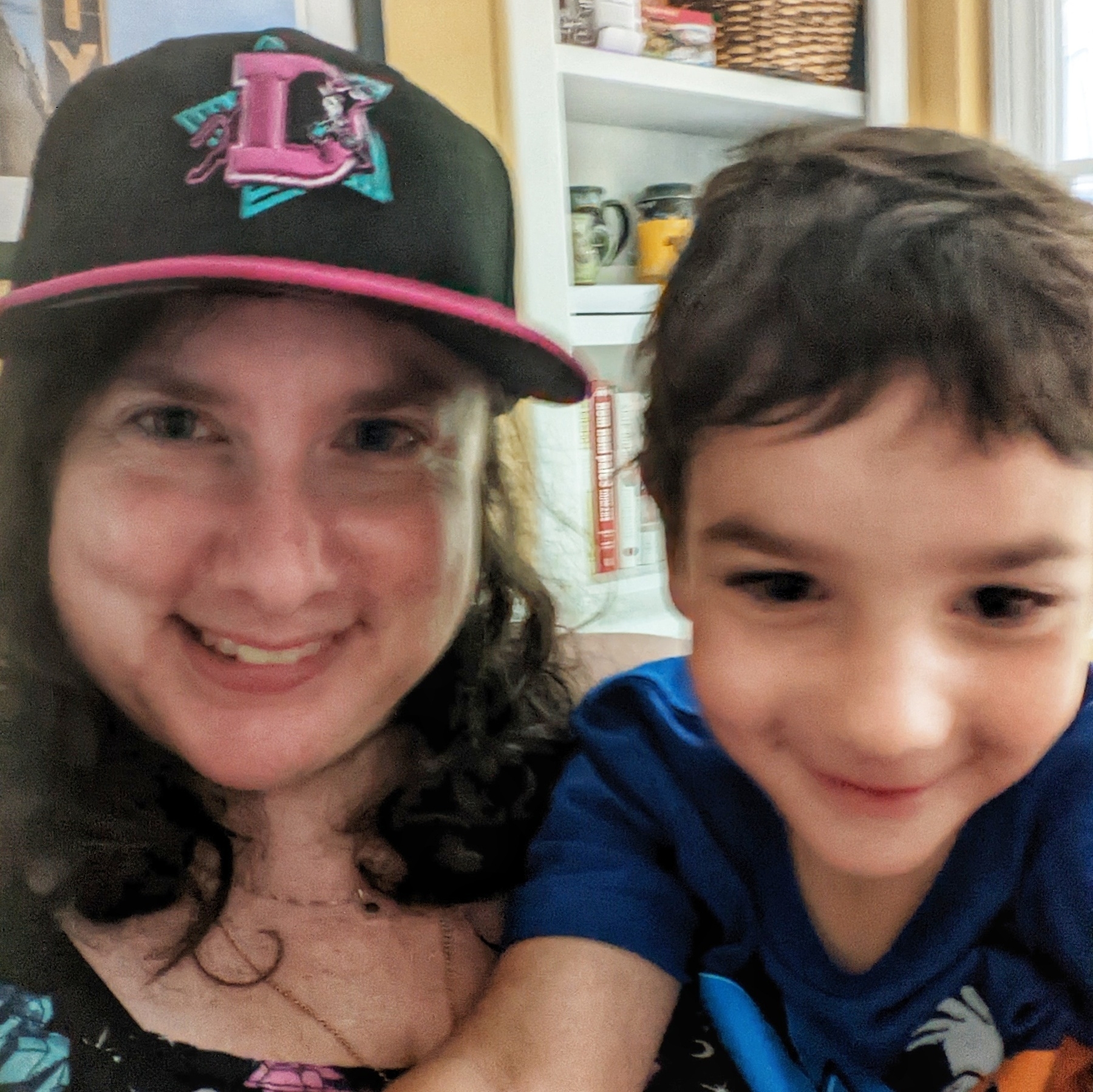 A white woman and a white child smile. They both have dark hair. The woman wears a black baseball cap with a pink brim and a logo with a bull jumping through a pink letter D, on the foreground of a turquoise star.