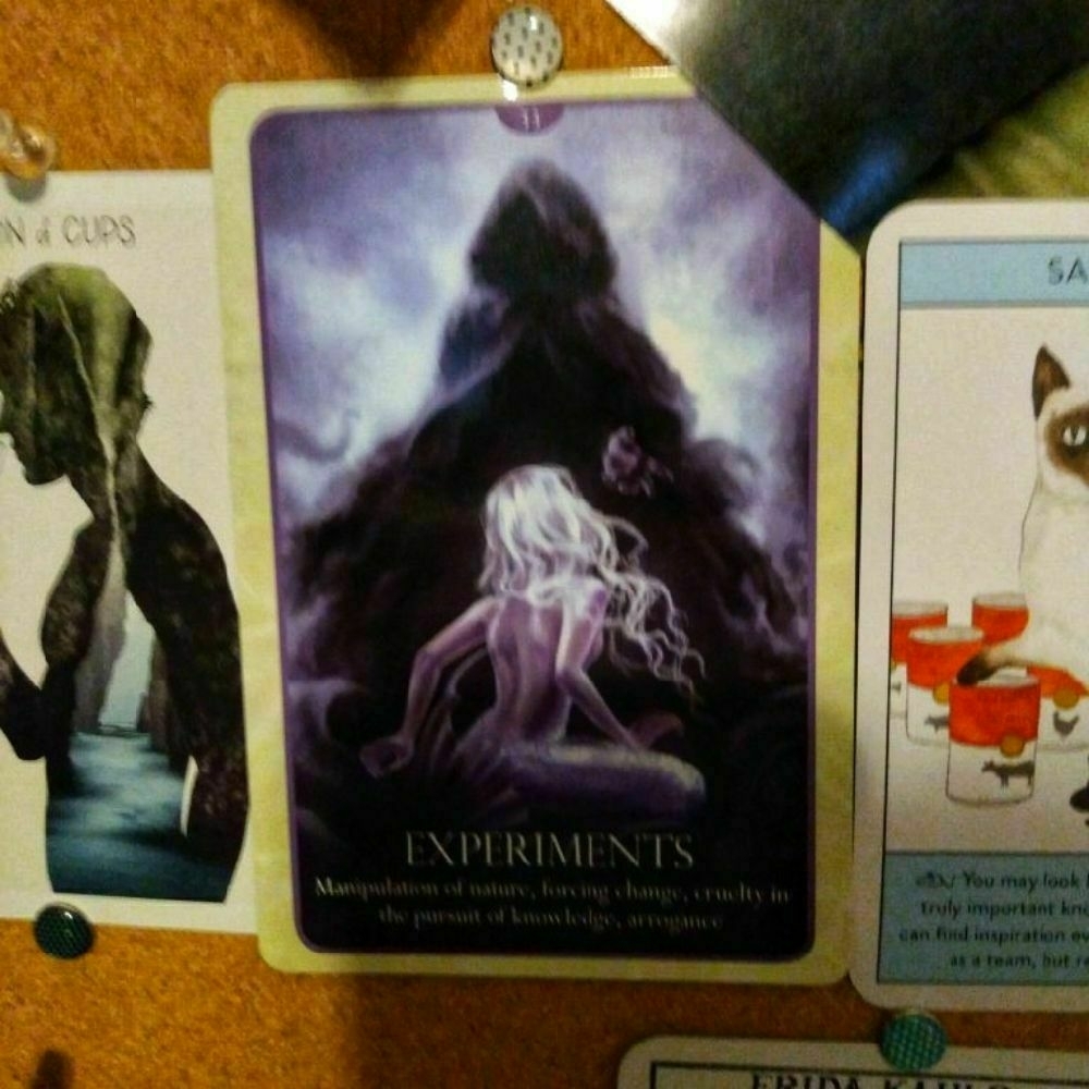 An oracle card depicts a silvery mermaid looking up at a shadowy cloaked figure stretching out their hand. Text on the card reads: "EXPERIMENTS Manipulation of nature, forcing change, cruelty in the pursuit of knowledge, arrogance"