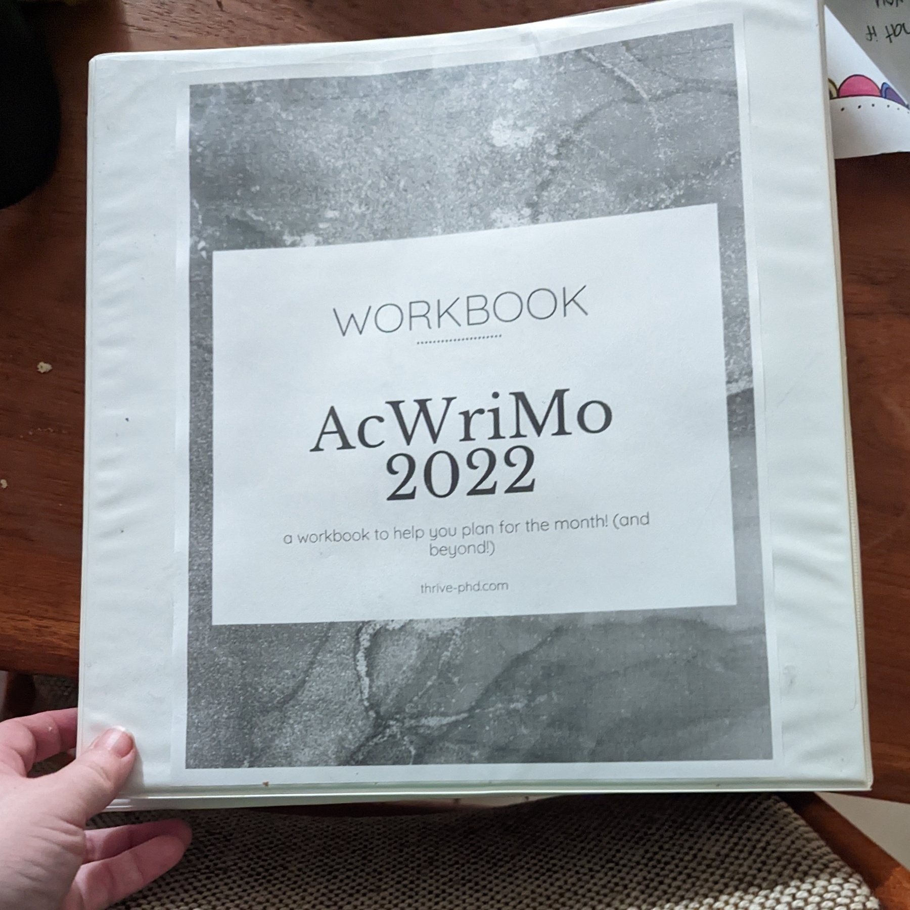 A hand holding a notebook with a cover that reads "Workbook AcWriMo 2022“