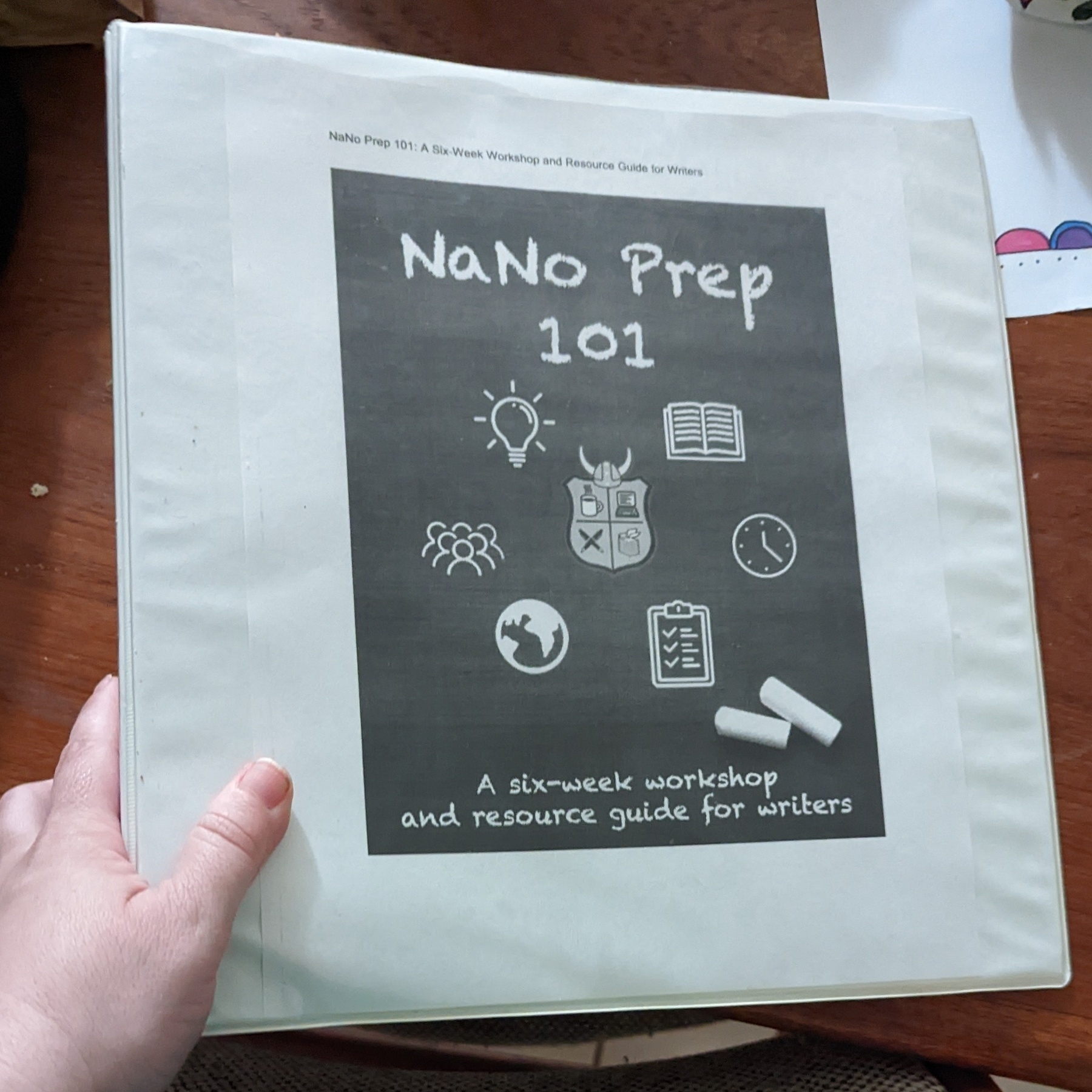 A hand holding a notebook with a cover that reads "NaNo Prep 101.“