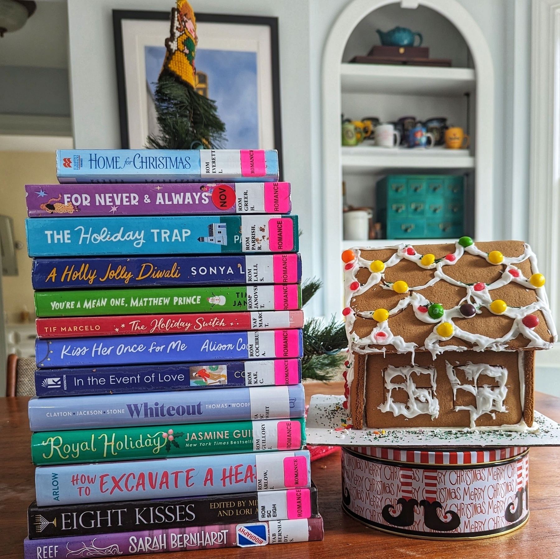 A stack of holiday romance novels sitting on top of a biography of Sarah Bernhardt, next to a gingerbread house that's sitting on top of a holiday tin.
